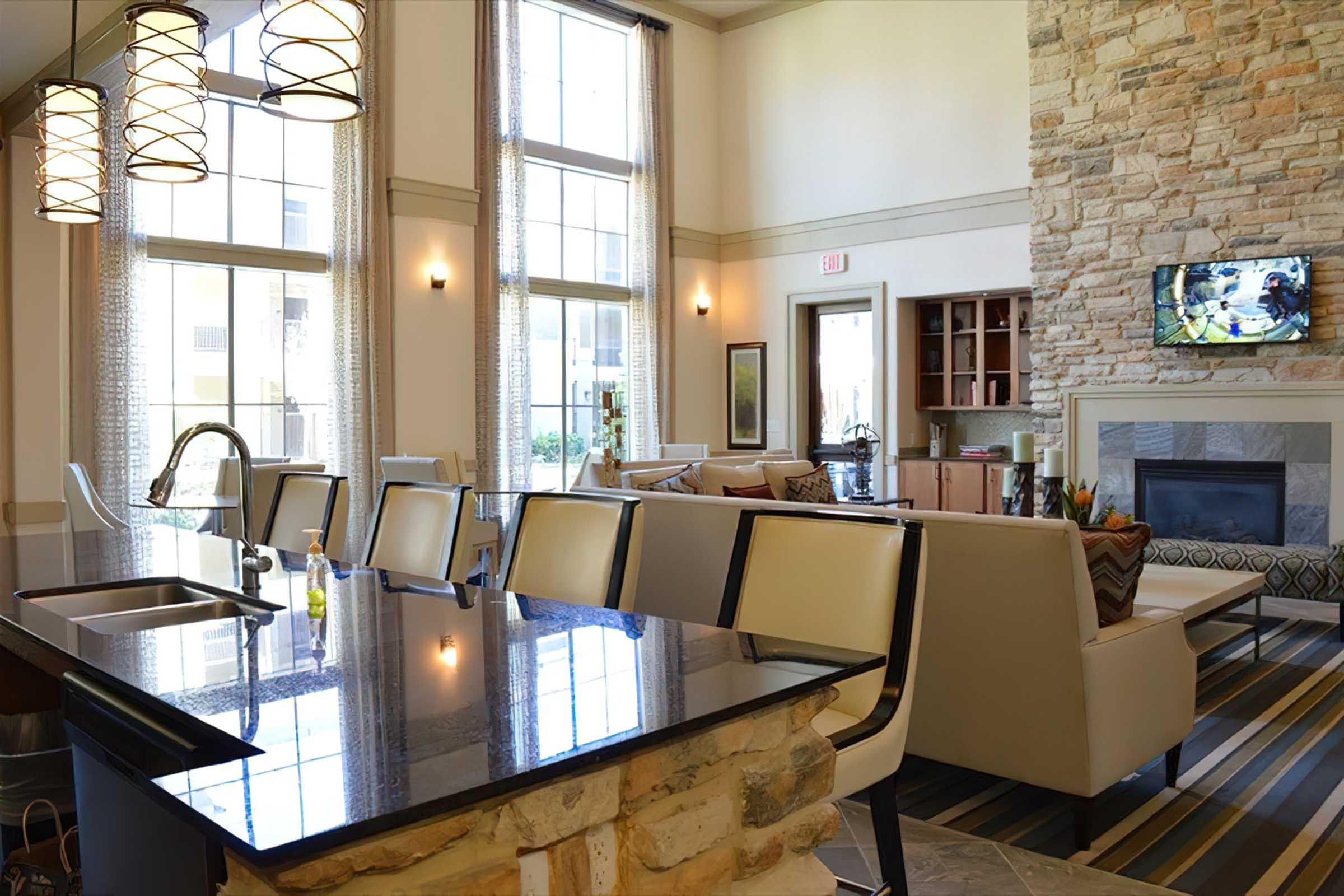 A modern lounge area featuring large windows with natural light, a stylish kitchen island with a dark countertop, and comfortable seating arrangements. The decor includes stone walls, striped rugs, and contemporary lighting fixtures, creating an inviting and elegant atmosphere. A TV is visible on the wall.