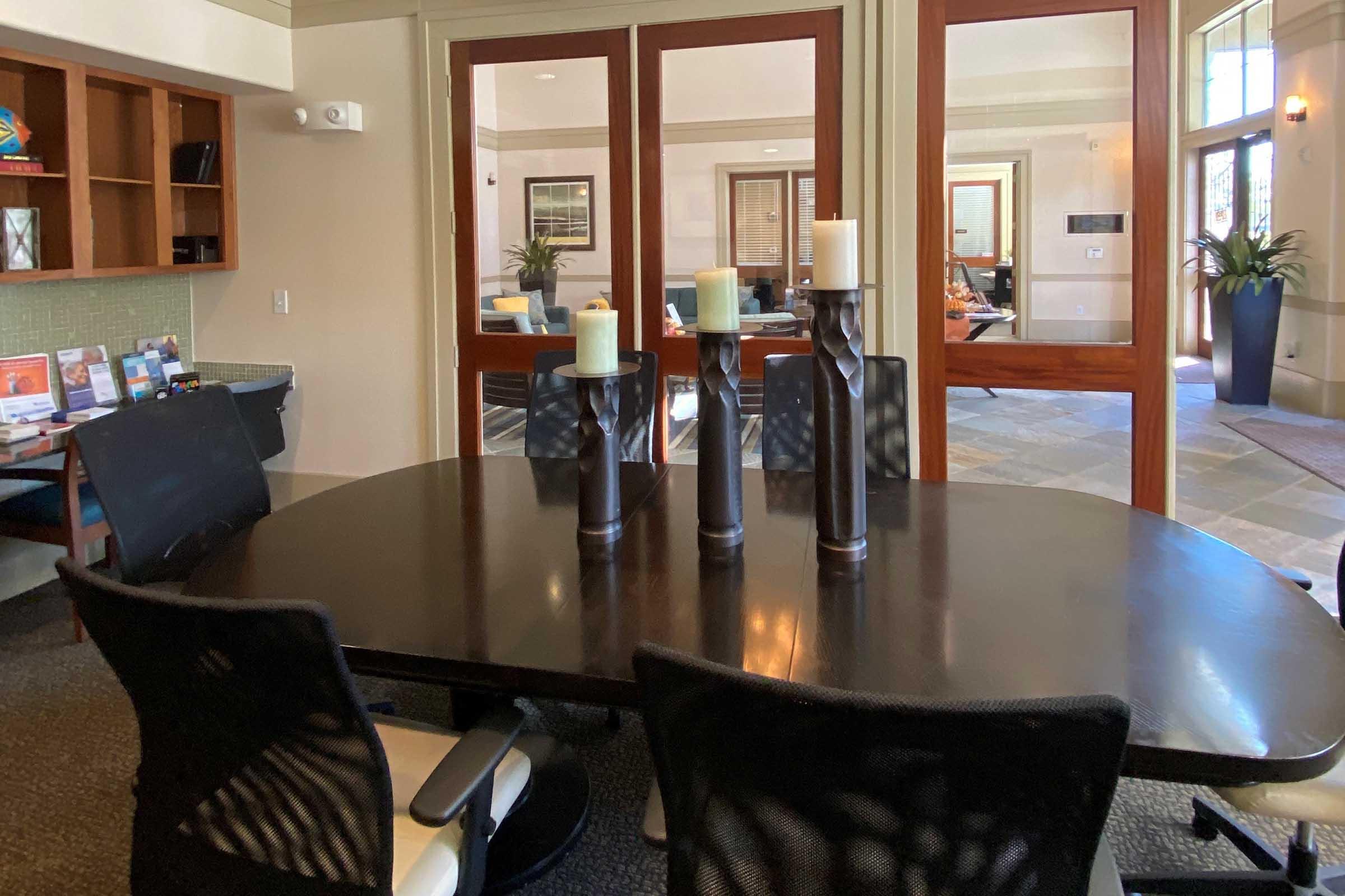 A modern conference room featuring a dark wooden table with three tall candle holders in the center, surrounded by black office chairs. Natural light enters through large glass doors, and a neatly organized workspace is visible in the background, along with decorative plants.