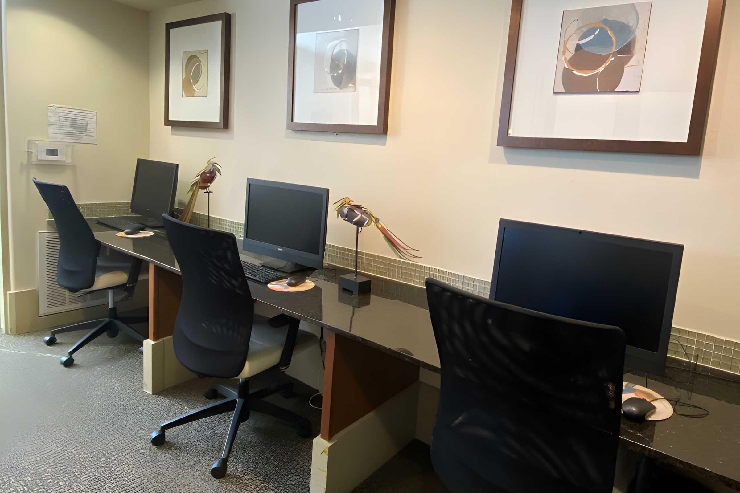 A workspace featuring four ergonomic chairs at desks with computers. Behind the desks are framed abstract artworks on the wall. There are two decorative lamps on the desks, and the floor has a textured design. The setting appears modern and functional, ideal for computer use.