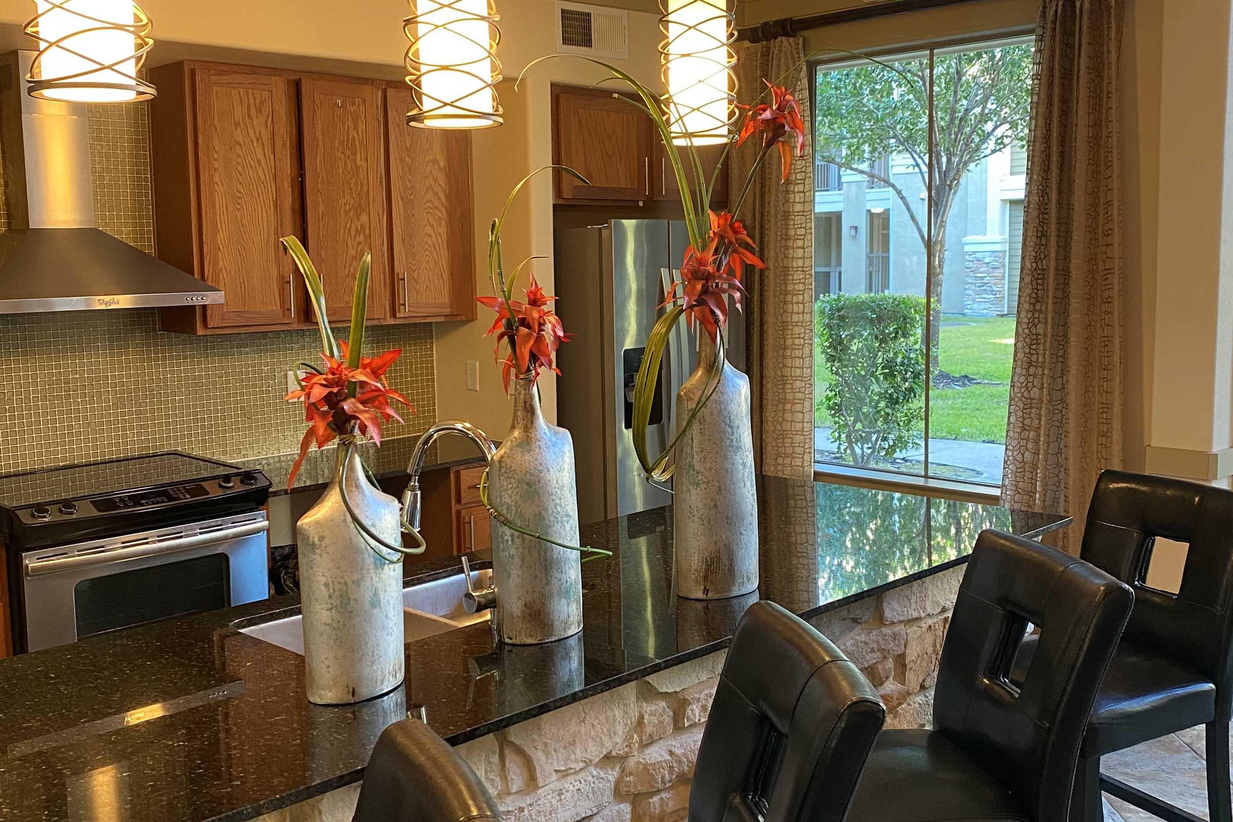 Modern kitchen interior featuring three decorative vases with red flowers, situated on a black granite countertop. Light wooden cabinets and stainless steel appliances are visible in the background, along with a window showcasing green outdoor scenery.