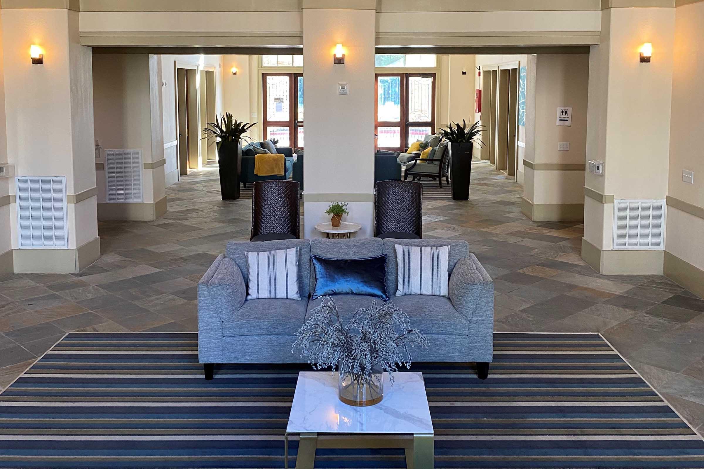 A modern lobby area featuring a gray couch with decorative pillows, a coffee table with a small plant, and a striped area rug. The space is well-lit with potted plants and seating in the background, creating a welcoming atmosphere.