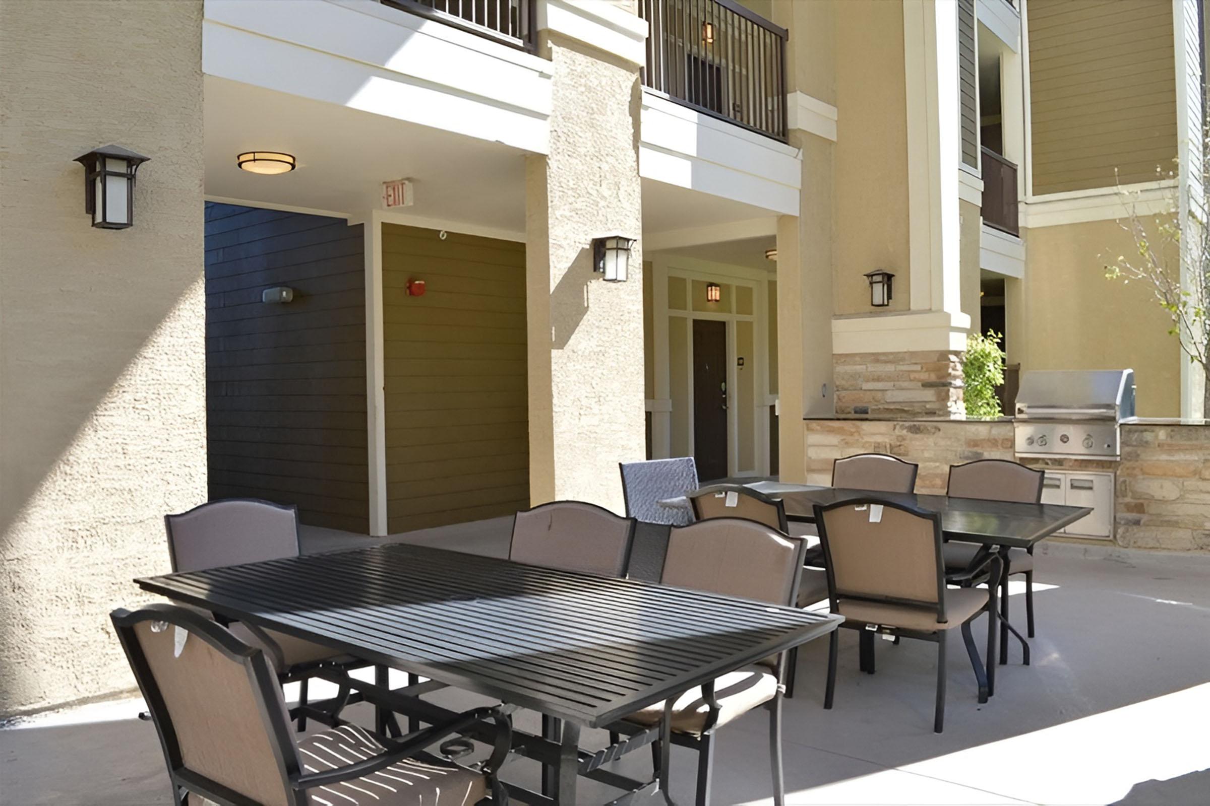 Outdoor dining area featuring multiple tables and chairs, overlooking a well-lit patio space of an apartment complex. There are grill facilities nearby, and the architecture includes large windows and a contemporary design with neutral colors.