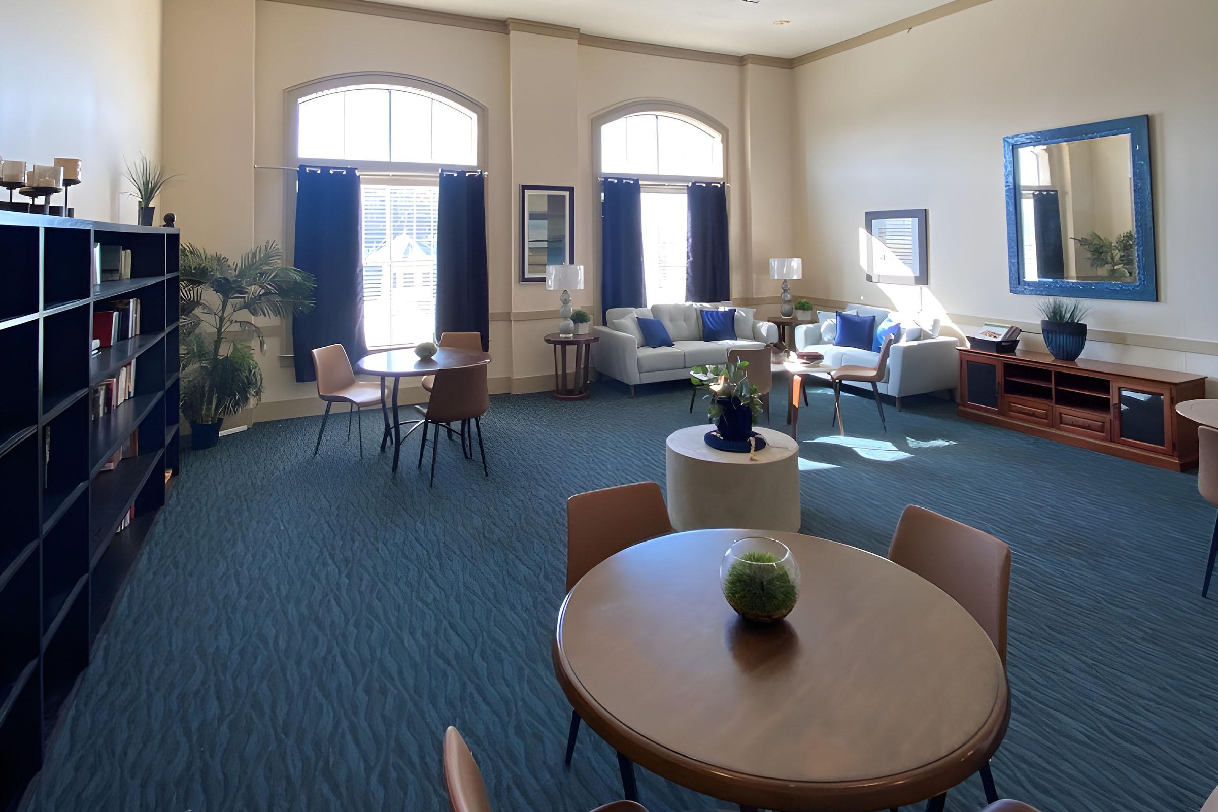 A spacious and well-lit lounge area featuring two round tables with chairs, a comfortable seating arrangement with cushions, and a decorative mirror on the wall. Large windows provide natural light, and there are plants placed around for added decor. The color scheme includes blues and neutral tones.