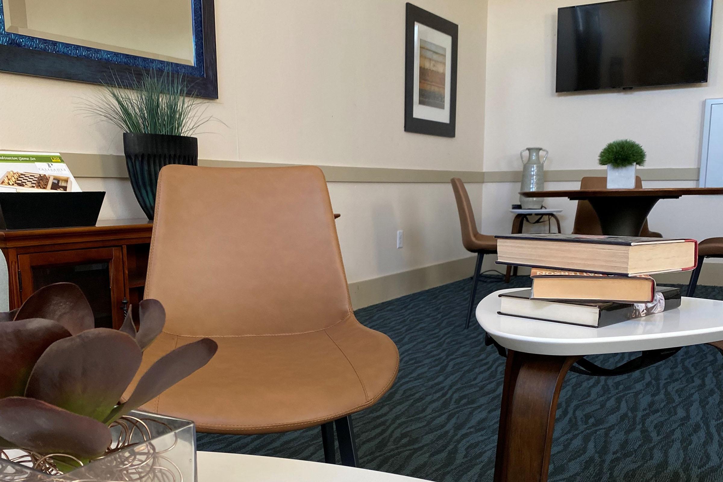 A cozy waiting area featuring a brown chair, a small table with stacked books, and a decorative plant. The room is decorated with a mirror and artwork, with a television mounted on the wall, creating a modern and inviting atmosphere.