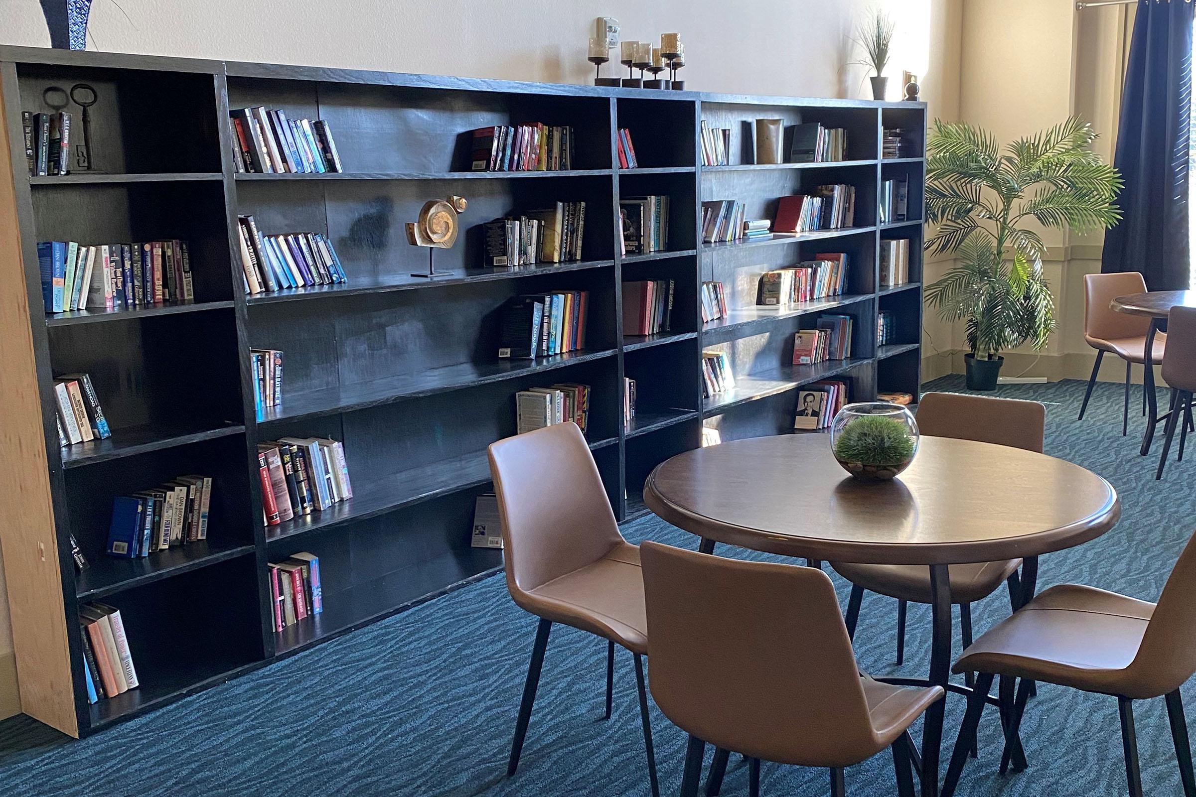 A cozy reading area featuring a large bookshelf filled with various books. In front of the bookshelf, there is a round table with a small decorative plant and several chairs arranged around it. The space is well-lit and has a modern design with a touch of greenery.