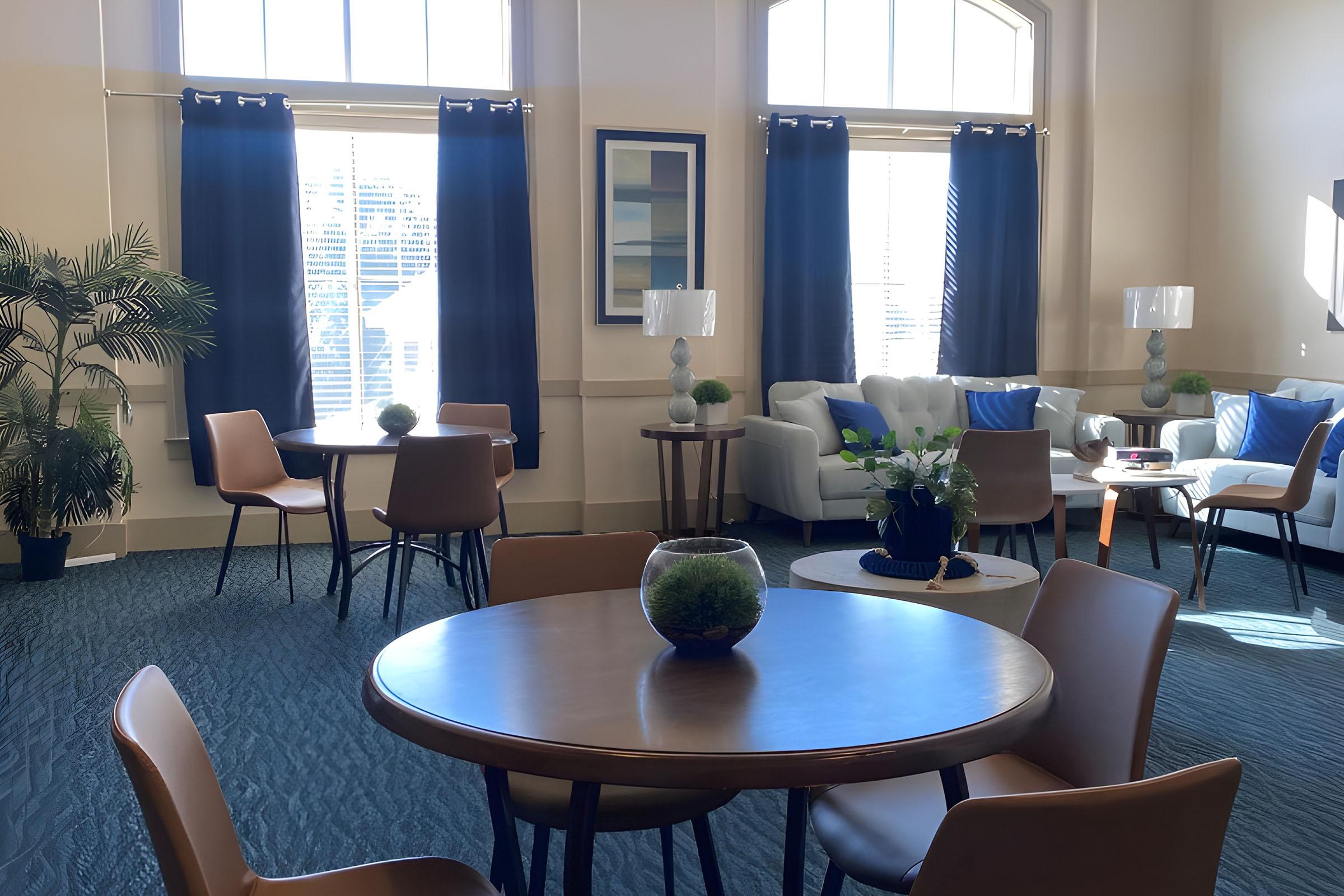 A bright and modern interior space featuring two tables with chairs, a cozy seating area with white couches and decorative lamps, potted plants, and large windows with dark curtains. The carpet is a light blue, creating a serene atmosphere.