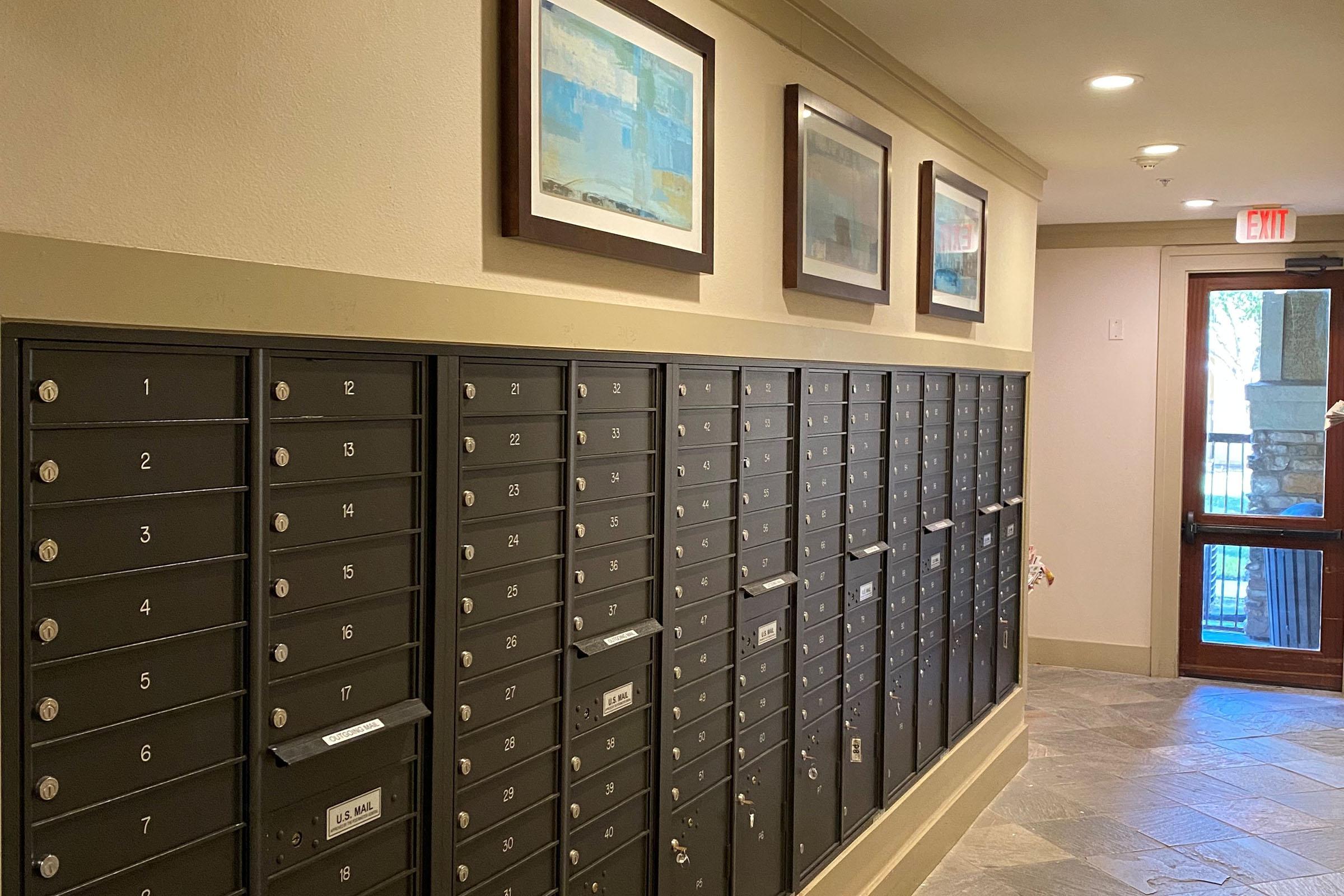 A hallway featuring a row of dark mailbox units numbered 1 to 26. Above the mailboxes are three framed abstract paintings. The floor is tiled, and there is a wooden door at the end of the hallway leading outside. Soft lighting enhances the modern design of the space.