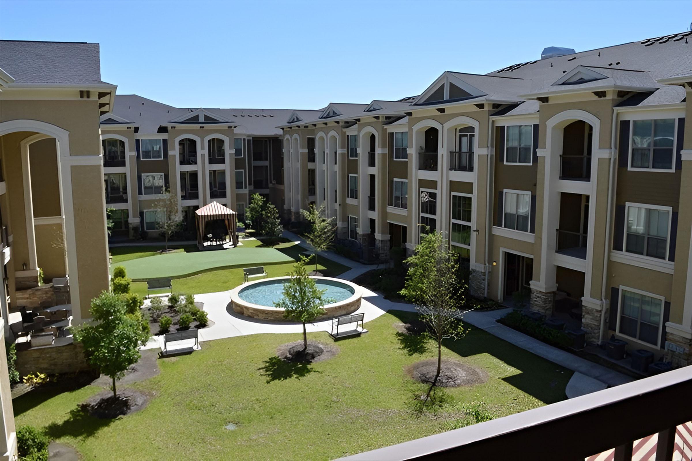 View of a landscaped courtyard in a residential complex featuring green lawns, a central fountain, benches, and a gazebo surrounded by multi-story apartment buildings under a clear blue sky.