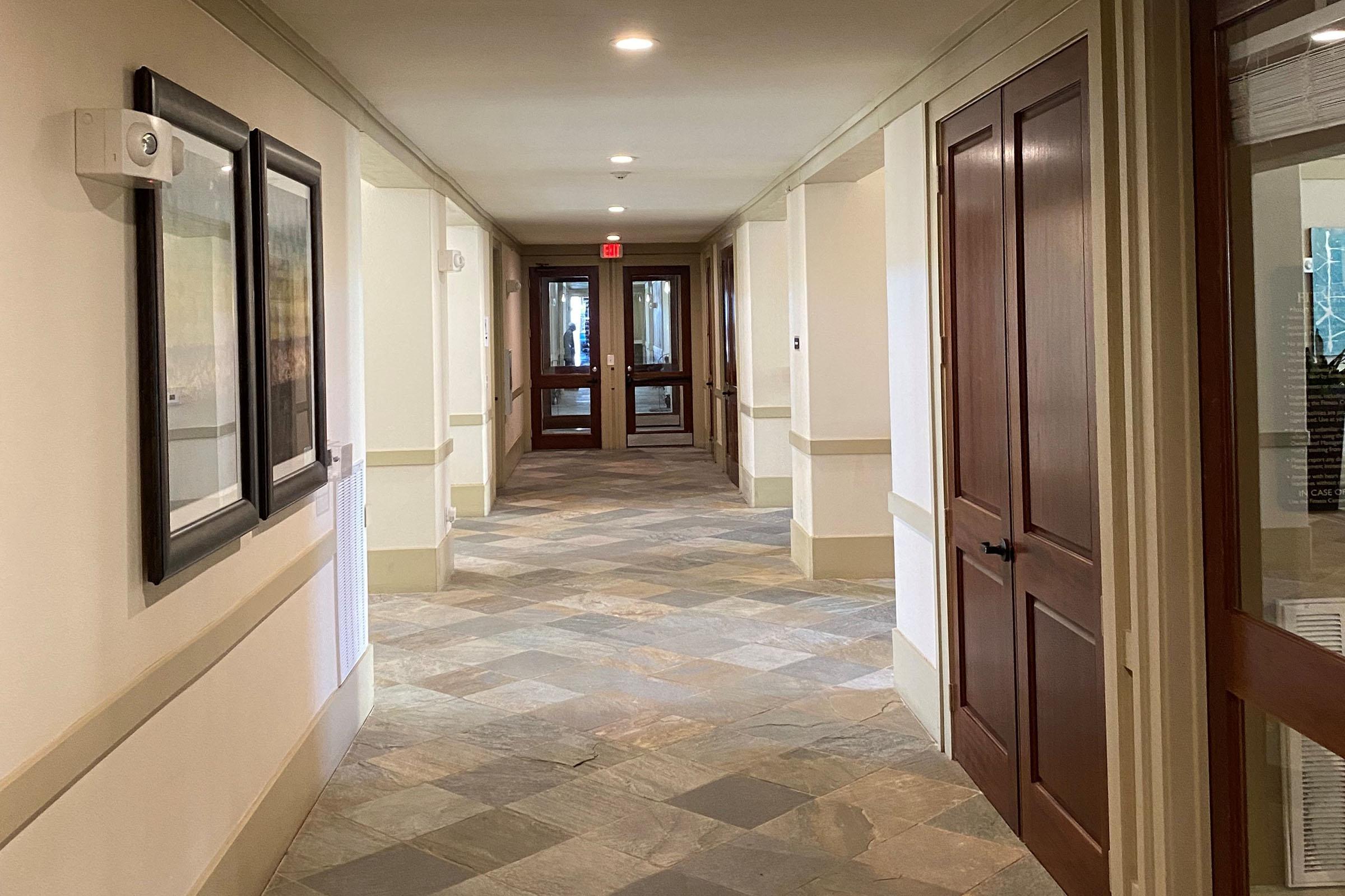 A well-lit corridor featuring tiled flooring and walls painted in neutral tones. On one side, there are framed artworks and doors, while at the end, double glass doors lead outside. The space has a clean and modern aesthetic, with natural light illuminating the hallway.