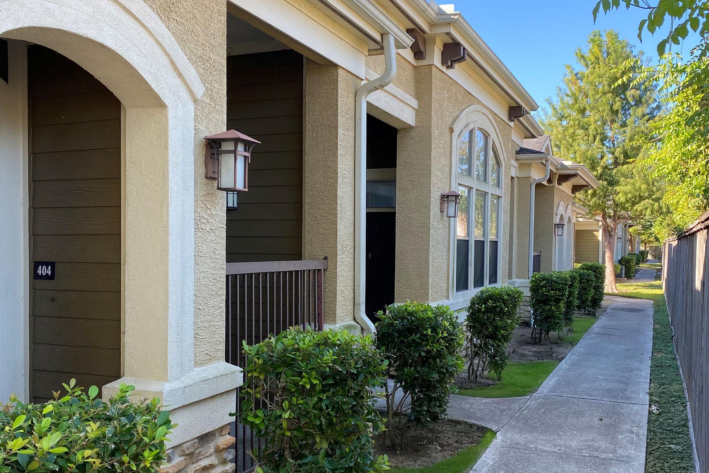 Row of beige apartment buildings with arched windows and porch lights, featuring small hedges and a concrete walkway. Lush green trees are visible in the background under a clear blue sky. The building numbers are partially visible, with the number "404" on one of the doors.