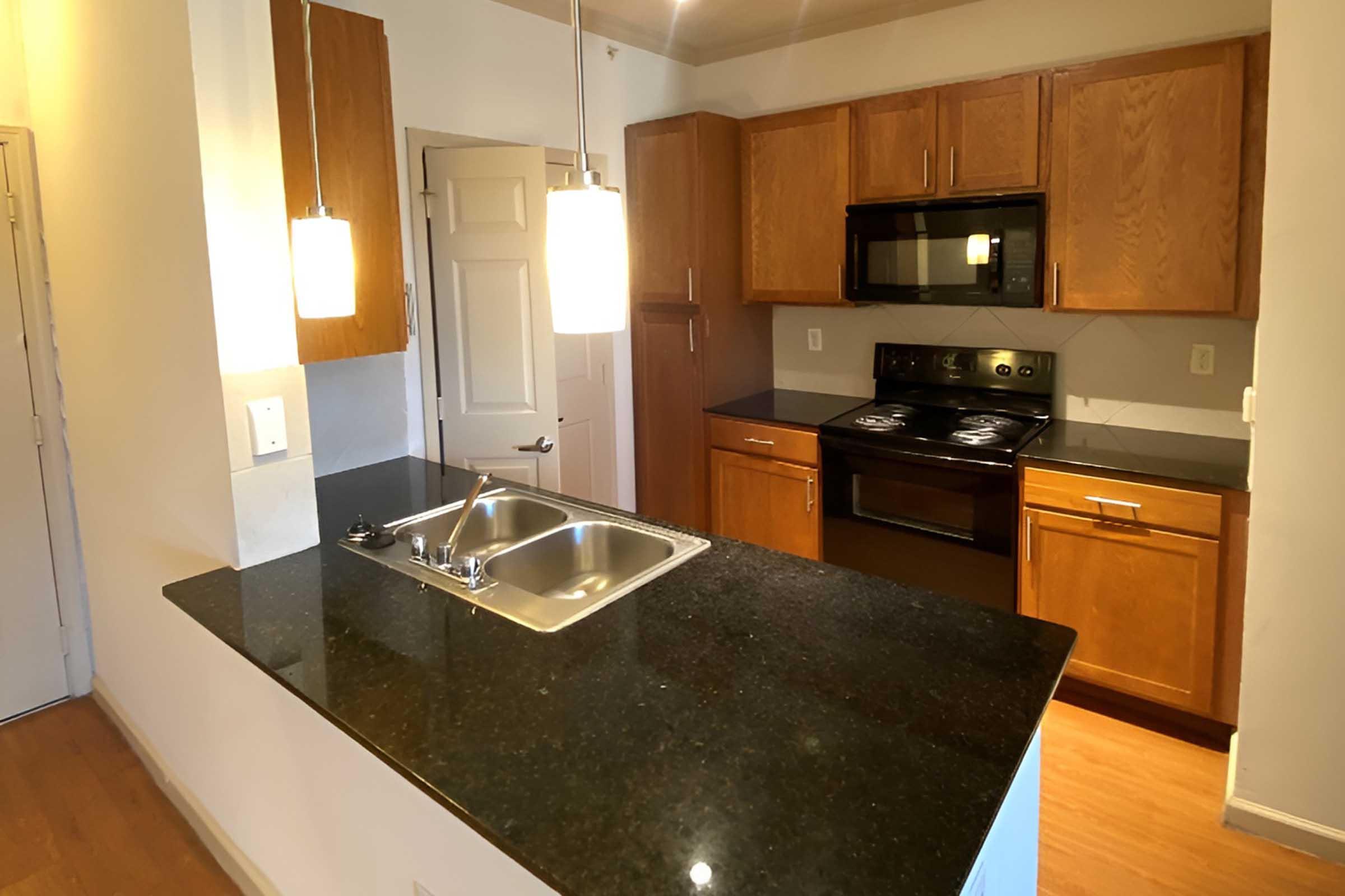 Modern kitchen featuring wooden cabinets, a black countertop, and stainless steel appliances. The kitchen includes a double sink, a stove, and a microwave. Pendant lights hang above the island, providing illumination. The space has a warm, inviting atmosphere with wooden flooring and neutral-colored walls.