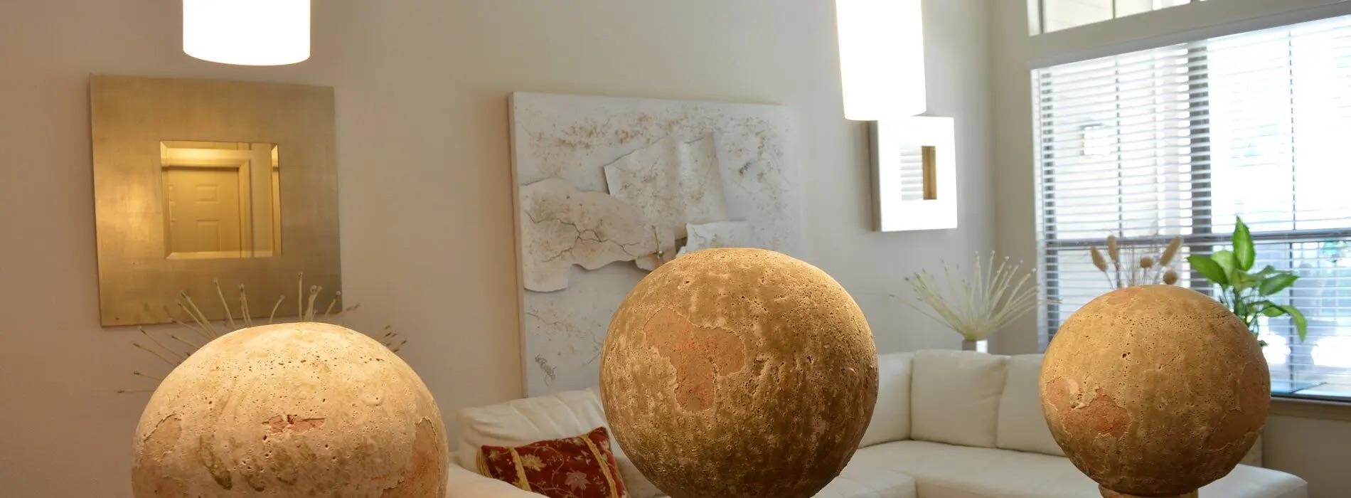 Three large, textured, spherical objects resembling planets are placed in a bright and modern living room. The room features a white couch, a decorative mirror, an abstract painting, and a houseplant by the window, creating a contrast between the natural textures of the spheres and the contemporary interior design.
