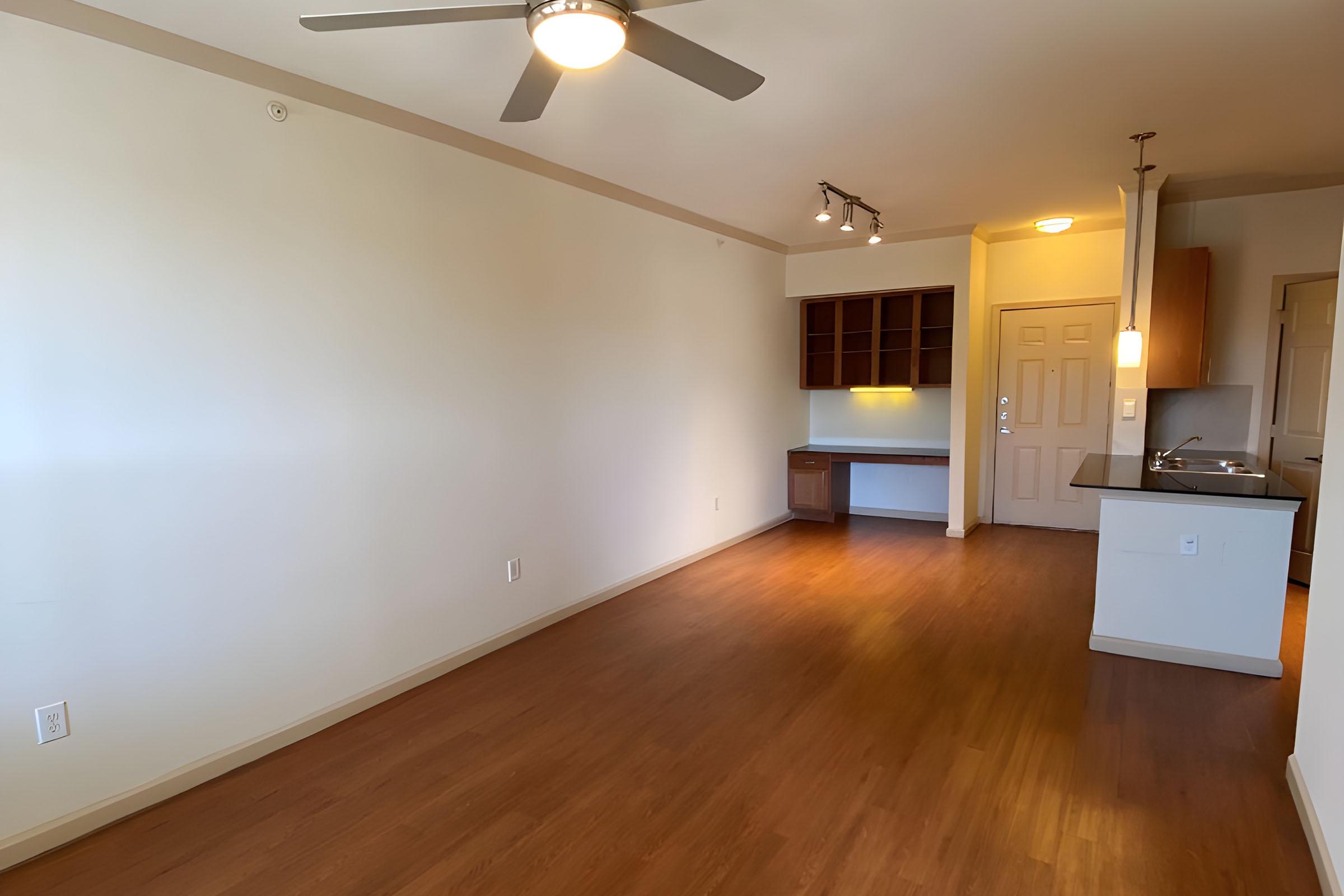 A spacious, empty apartment interior with light wood flooring. The room features a ceiling fan, a small kitchenette with cabinets, and a doorway leading to another space. Natural light fills the area, creating a bright and airy atmosphere.