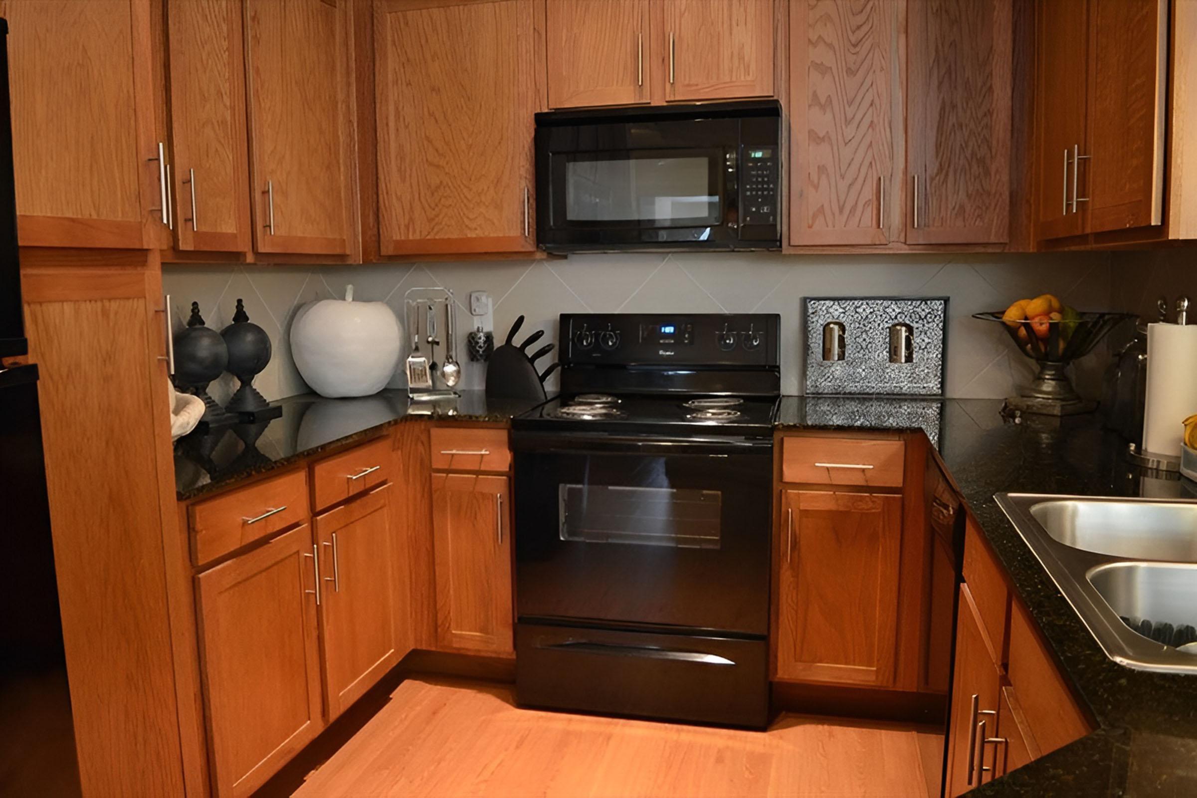 A modern kitchen featuring wooden cabinets, a black stove and microwave, and a countertop with a dark granite finish. Decorative items, including a white vase, sit beside a bowl of fruit. The kitchen has stainless steel appliances and a double sink, creating a functional and stylish space.