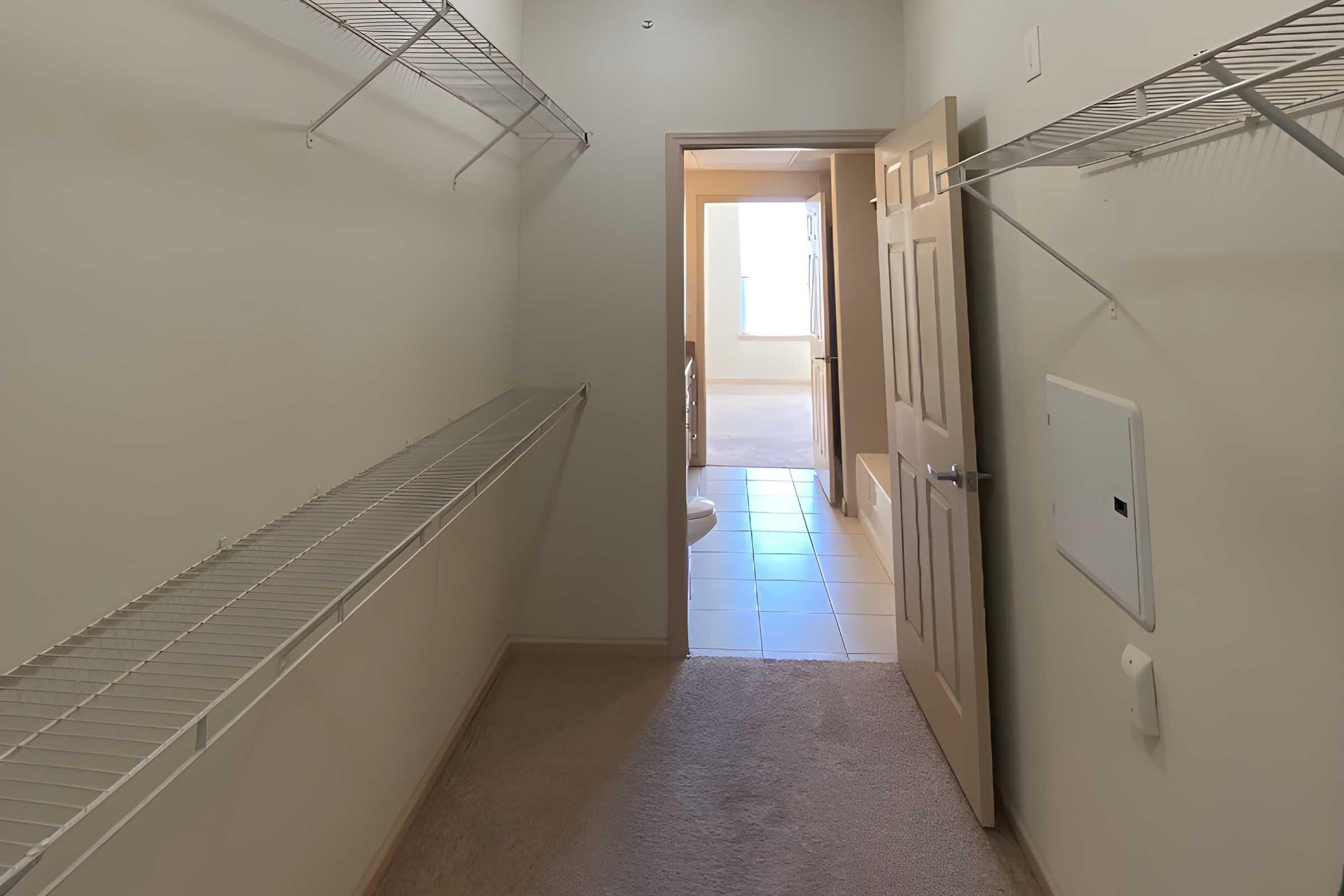A well-lit closet with wire shelving on both sides. The door is open, revealing a bathroom with tiled flooring and light-colored walls. The closet features beige carpet and a clean, organized appearance, creating a spacious feel.