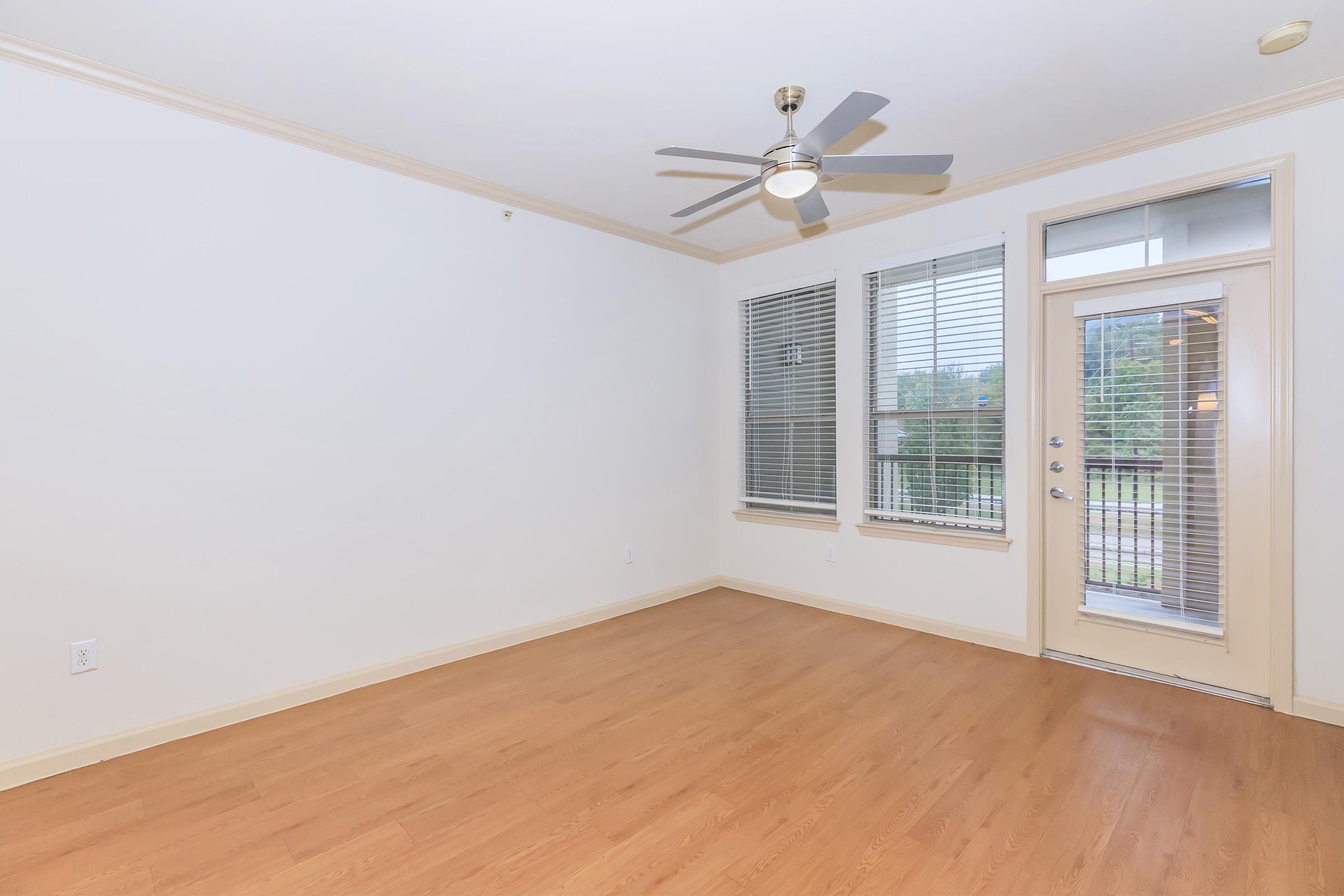 Spacious interior of a vacant room featuring light wood flooring, a ceiling fan, large windows with blinds, and a door leading outside. The walls are painted in neutral tones, creating a bright and airy atmosphere.