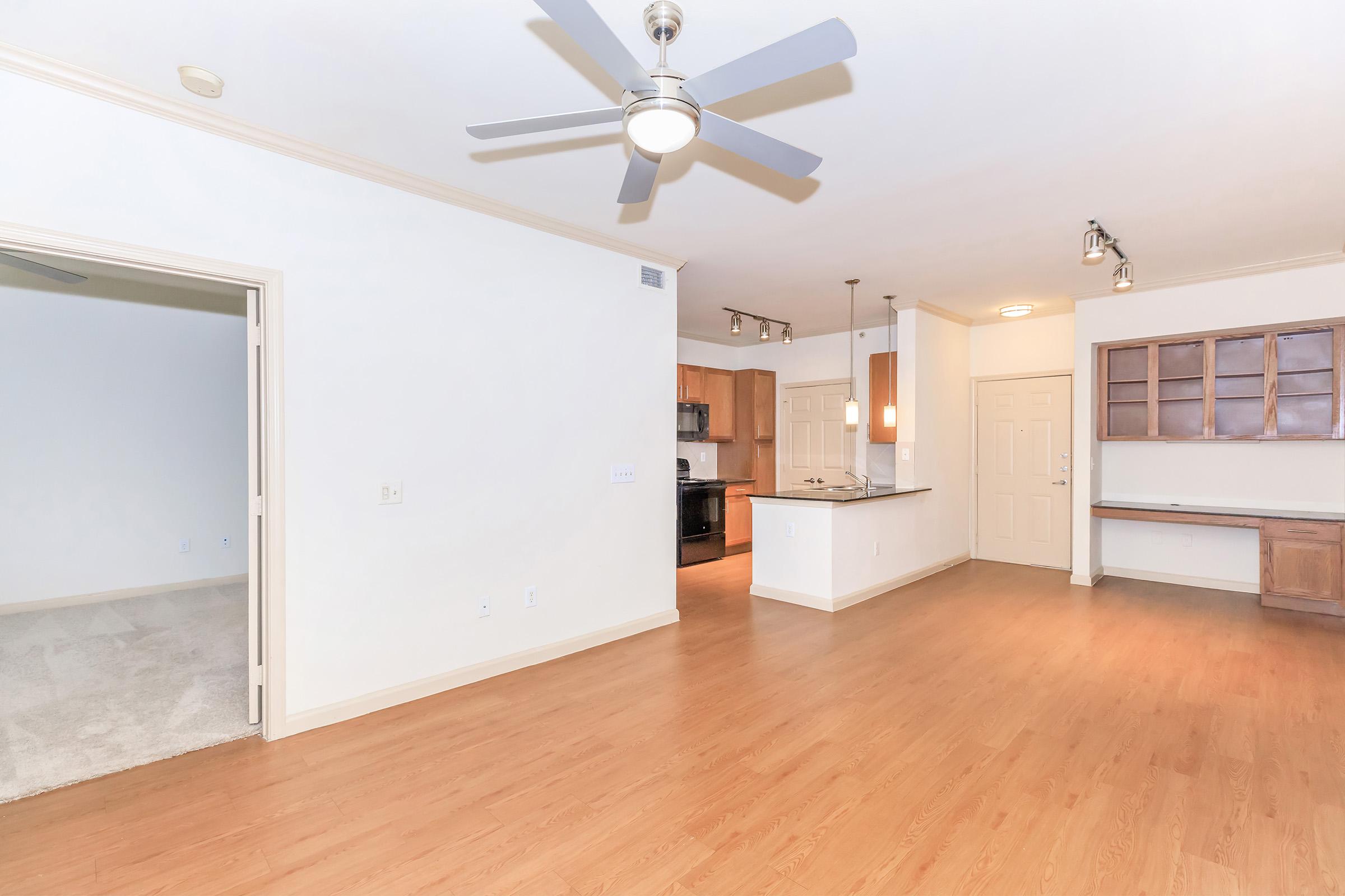 Bright and spacious living area featuring hardwood floors, a ceiling fan, and an open floor plan leading to the kitchen. The kitchen includes modern appliances and ample counter space. A doorway on the left opens to a separate room, while a built-in cabinet is visible along the right side.
