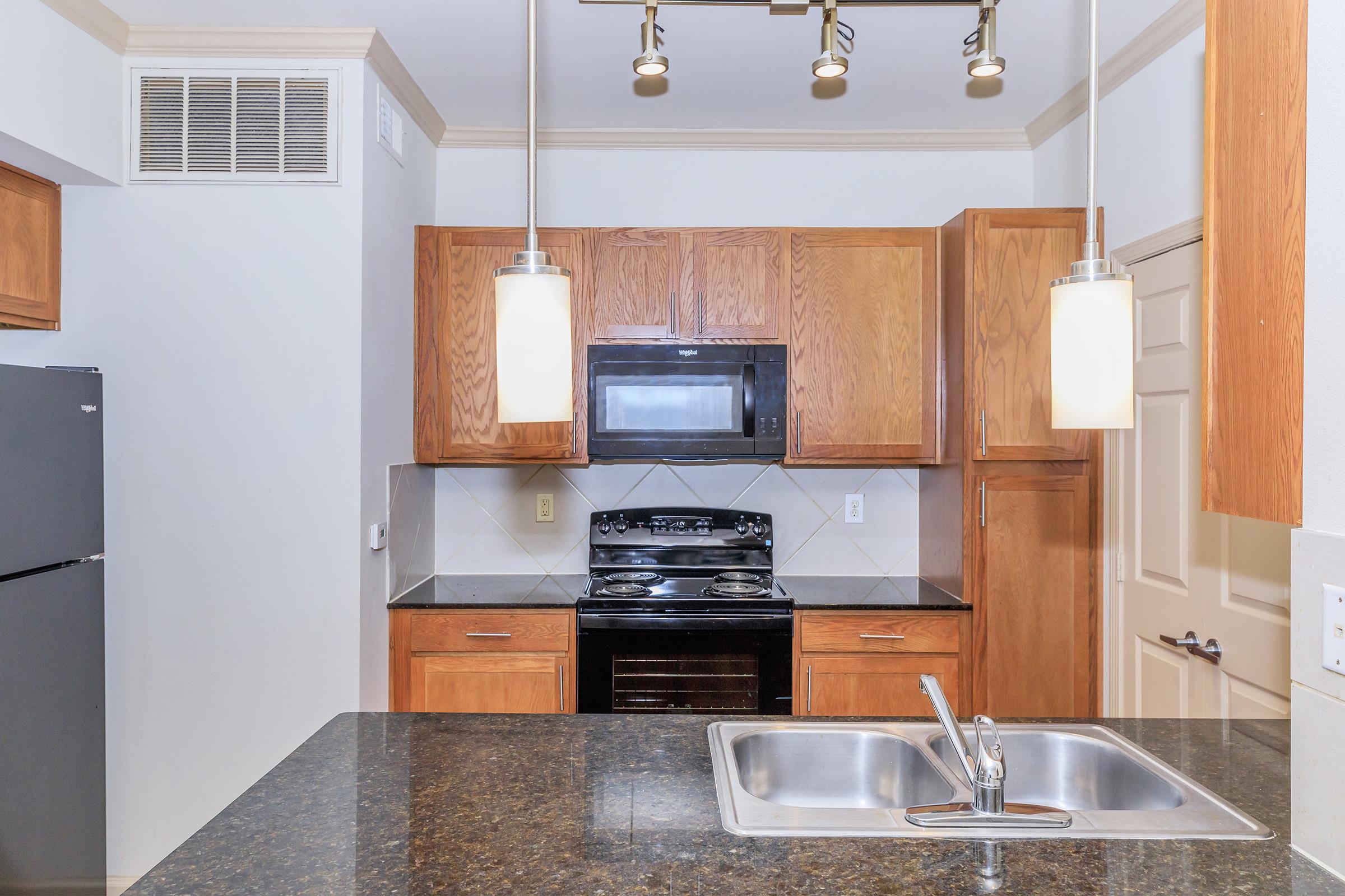 Modern kitchen featuring wooden cabinetry, black appliances including a microwave and stove, and a granite countertop with a double sink. There are pendant lights above the counter, and the overall space has a neutral color palette.