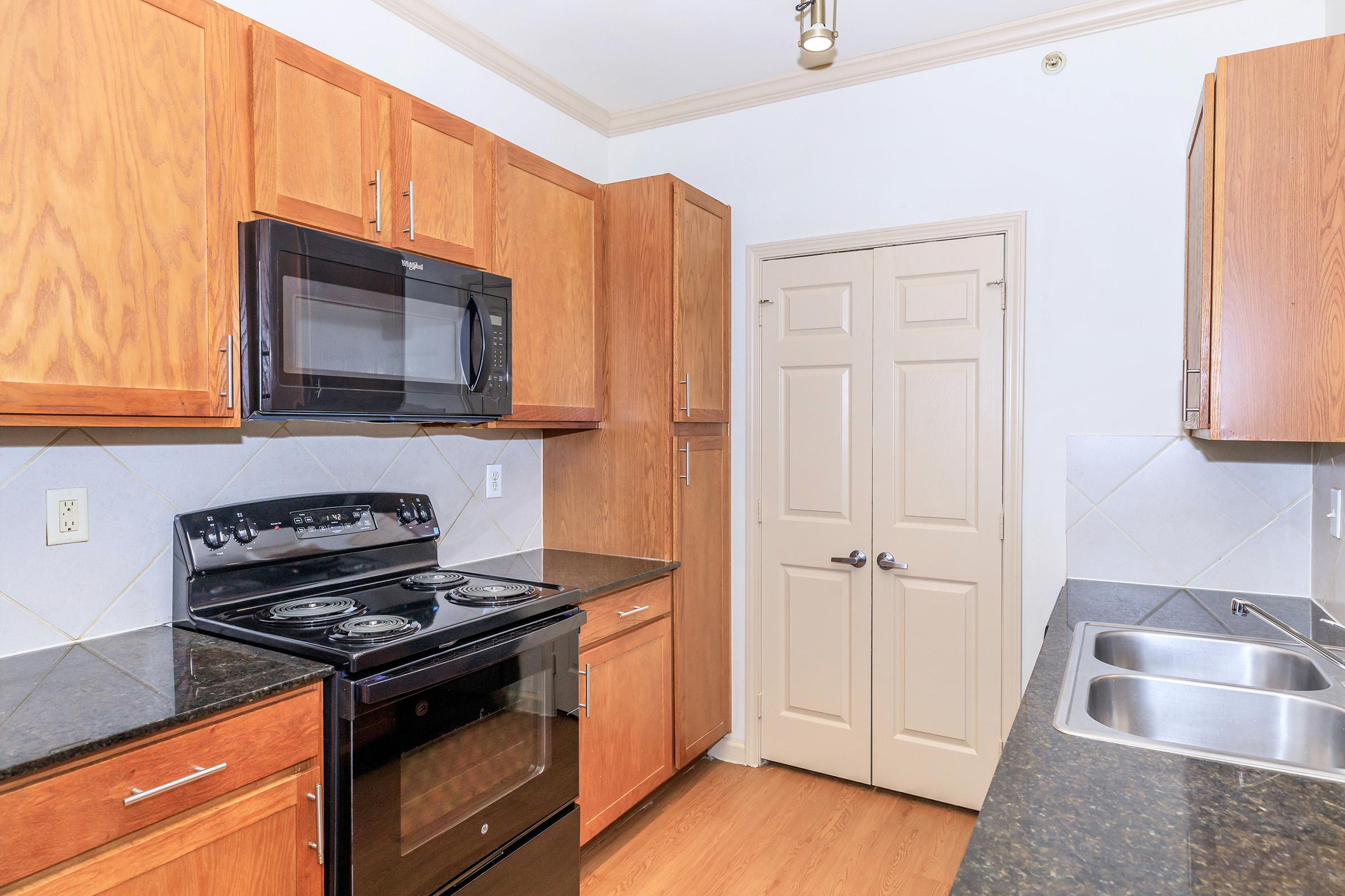 A modern kitchen featuring wooden cabinetry, a black stove and microwave, dark countertops, and a double sink. The walls are light-colored, and a door is visible, leading to another room. The flooring is a light wood laminate, creating a warm atmosphere.