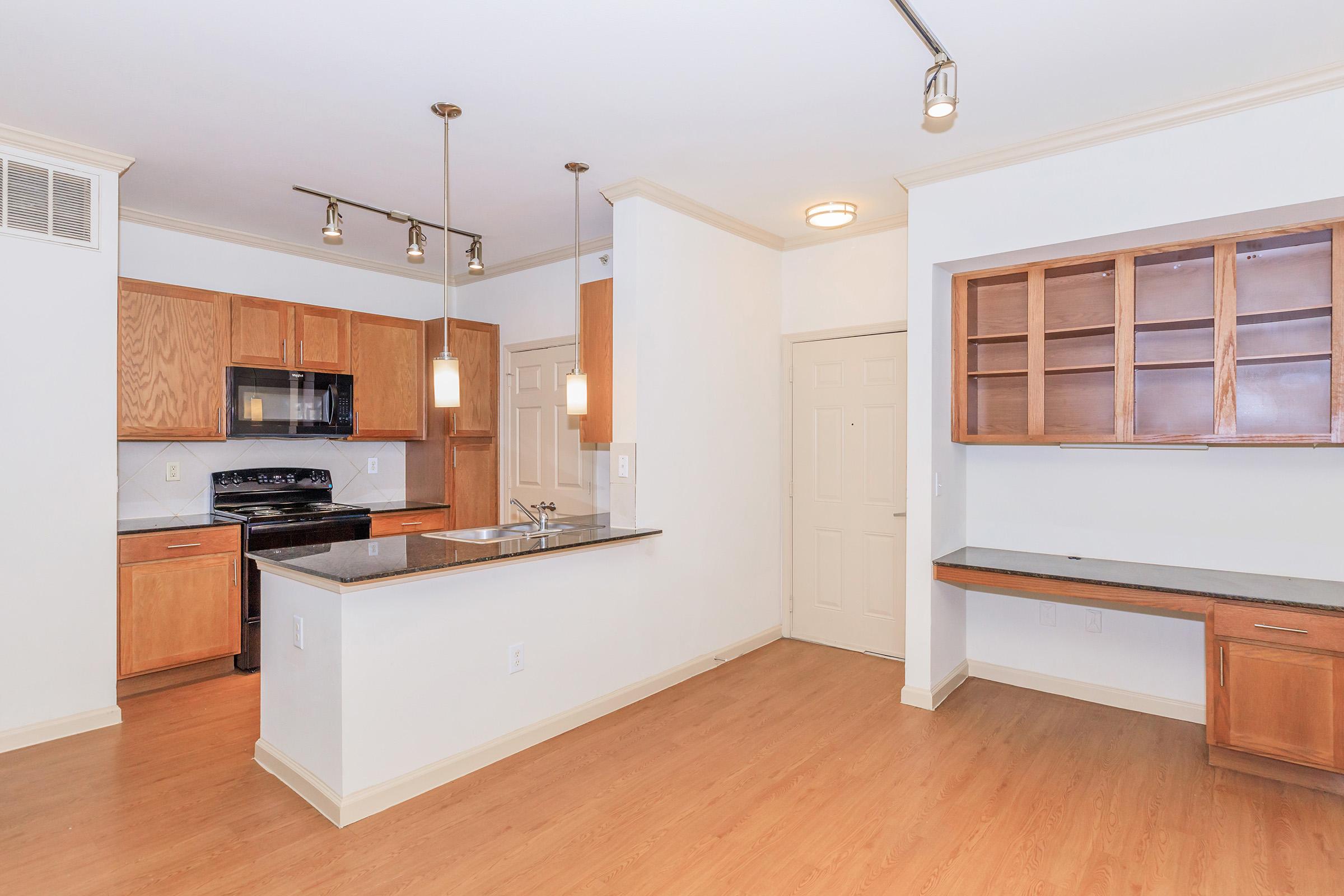 A modern kitchen with wooden cabinets, granite countertops, and stainless steel appliances. The open floor plan includes a living area with natural light. Pendant lighting hangs above the kitchen island, while a built-in shelf is visible on one wall, enhancing the spacious feel of the room.