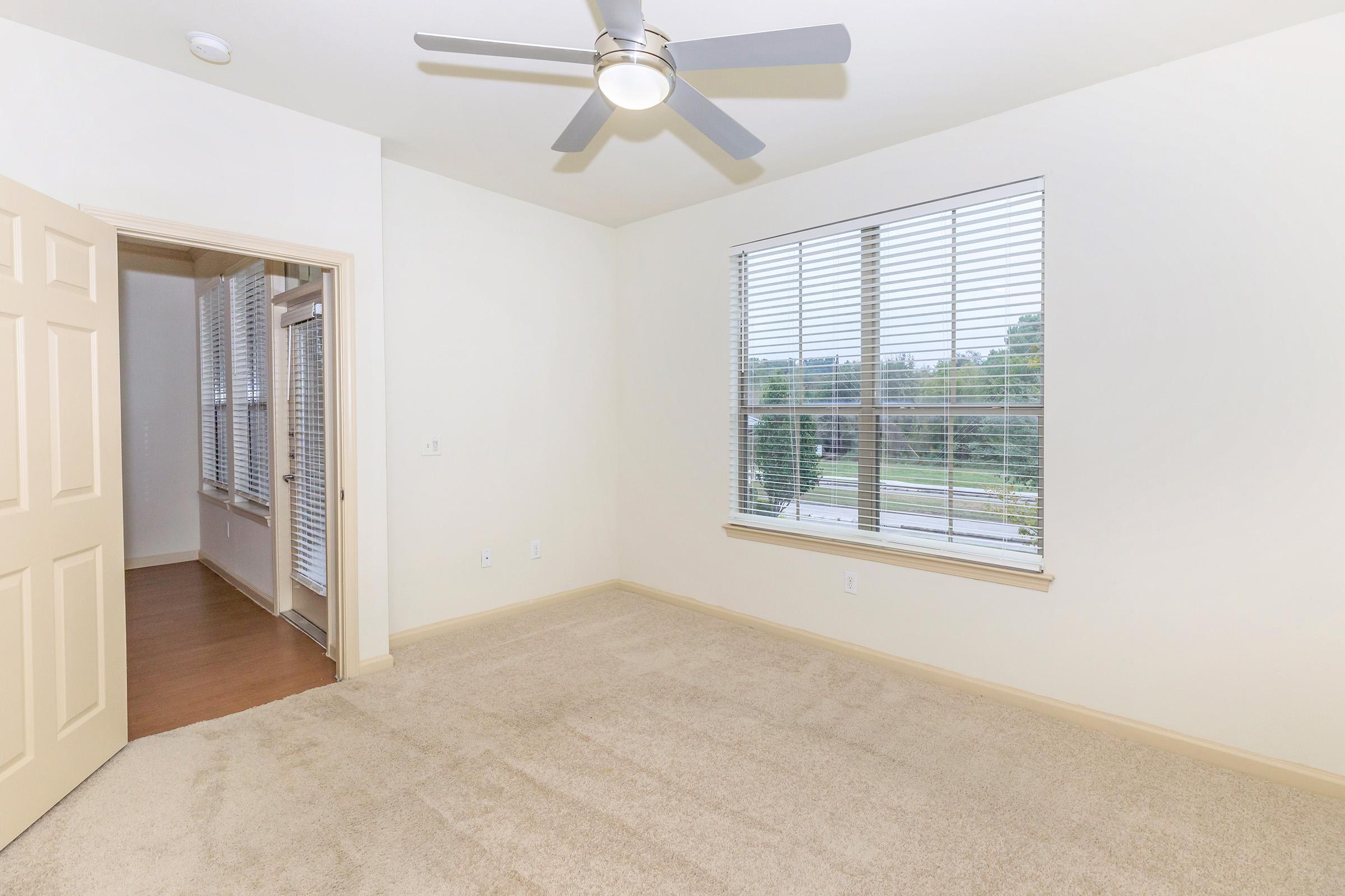 A well-lit, empty room featuring light-colored walls and carpet. There is a ceiling fan with three blades and a large window with blinds, allowing natural light to enter. A partially opened door leads to another area, creating a spacious feel. Outside the window, greenery is visible.