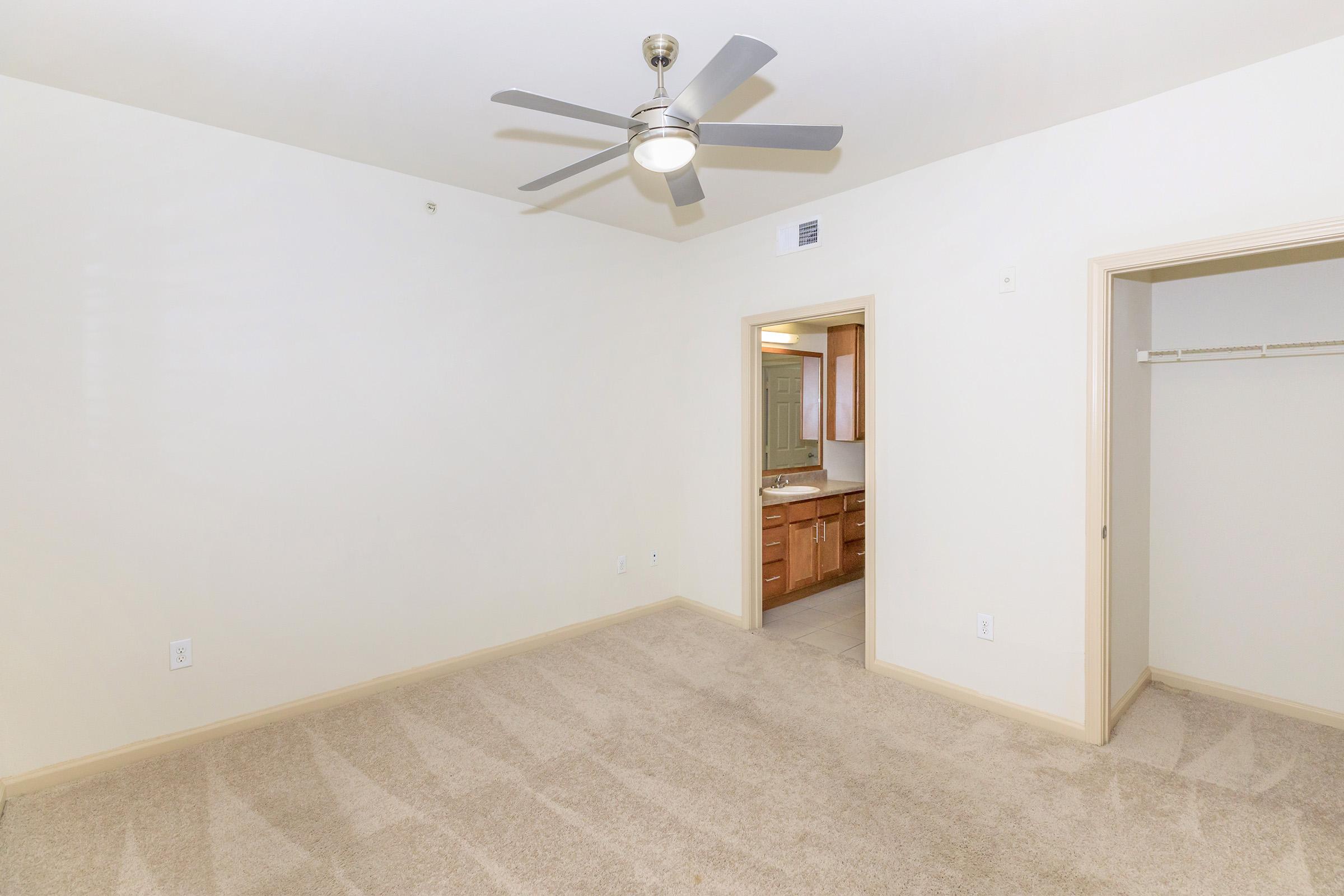 A spacious, lightly colored bedroom with beige carpet, featuring a ceiling fan. The room has two doorways: one leading to a bathroom with wooden cabinetry and the other to a closet. Natural light floods the space, creating a bright and inviting atmosphere.