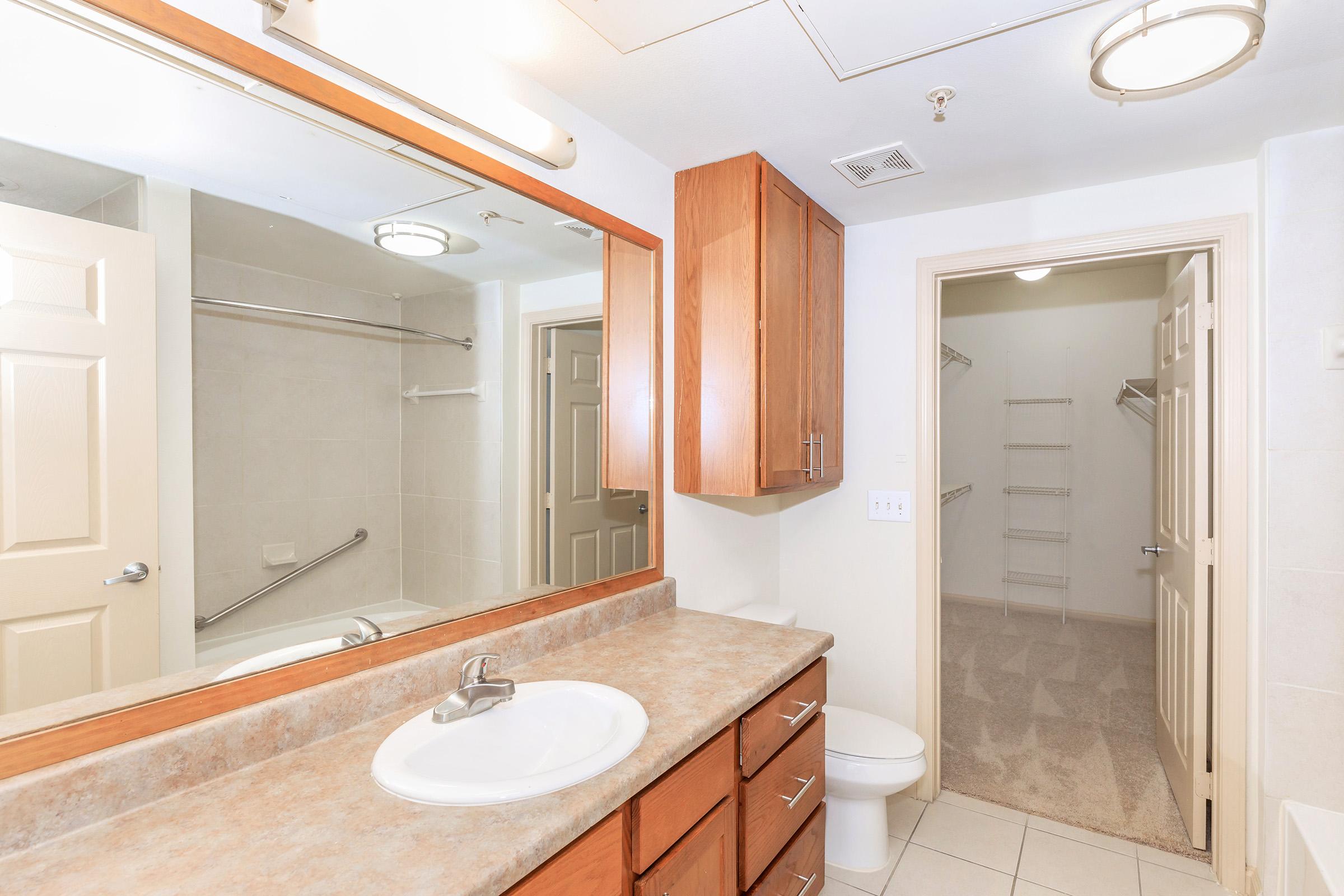 A modern bathroom featuring a large mirror above a countertop with a sink, wooden cabinetry, and a towel rack. In the background, there's an open door leading to a walk-in closet with shelving and a well-lit atmosphere. The space has neutral tile flooring and walls, creating a clean and spacious feel.