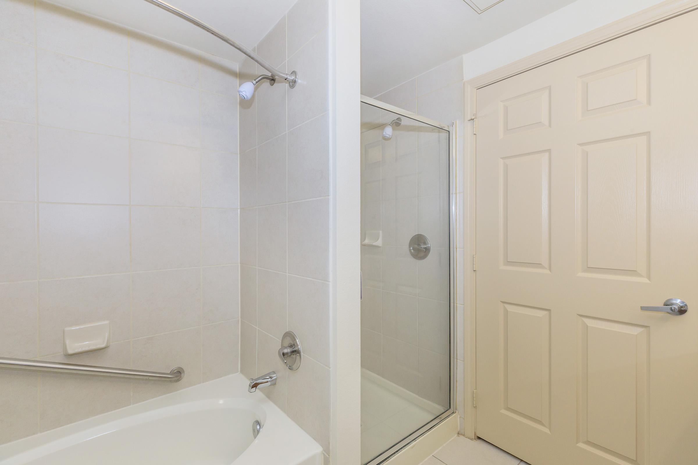 A clean bathroom featuring a bathtub with a shower head and grab bar, next to a glass shower enclosure. A beige door is visible on the right side, and the walls are tiled in light colors. The floor is tiled and the overall space is well-lit and modern.