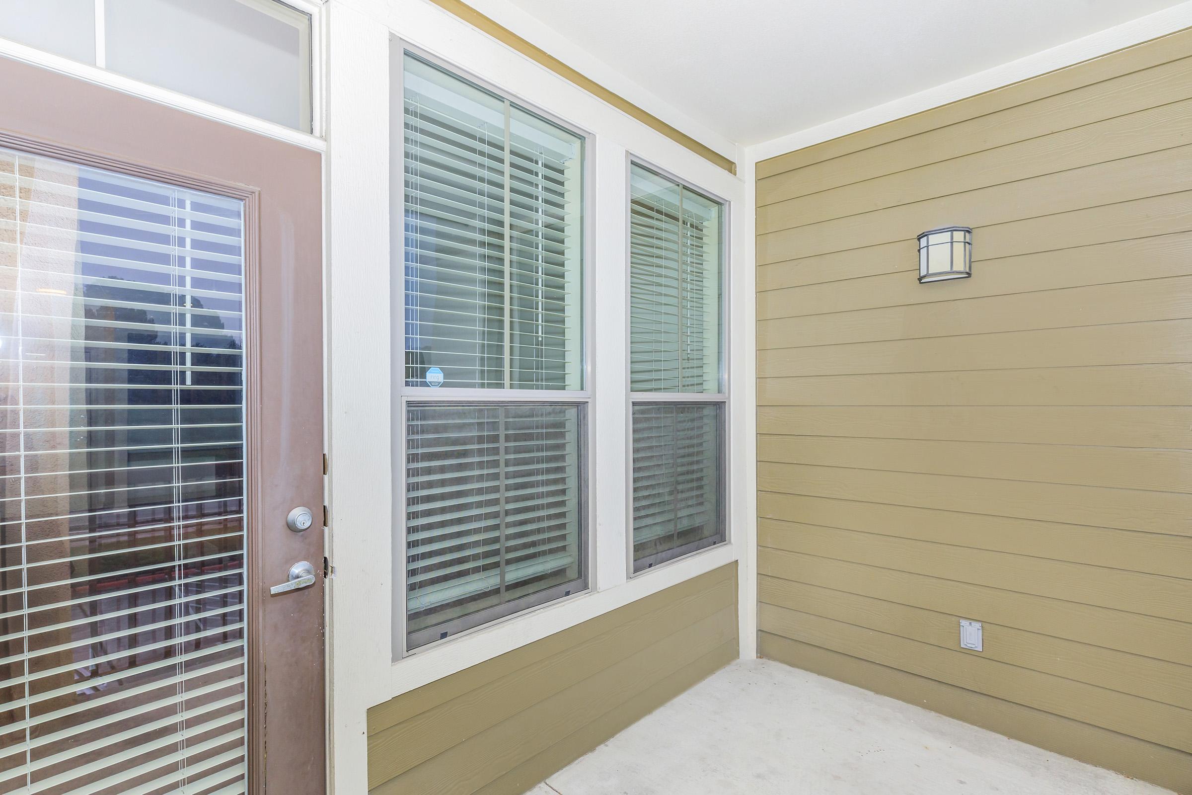 A modern entryway featuring a light brown exterior wall and large windows with white blinds. The front door has a modern handle and a glass element, while a coordinating light fixture is mounted on the wall. The space is well-lit, creating an inviting atmosphere.