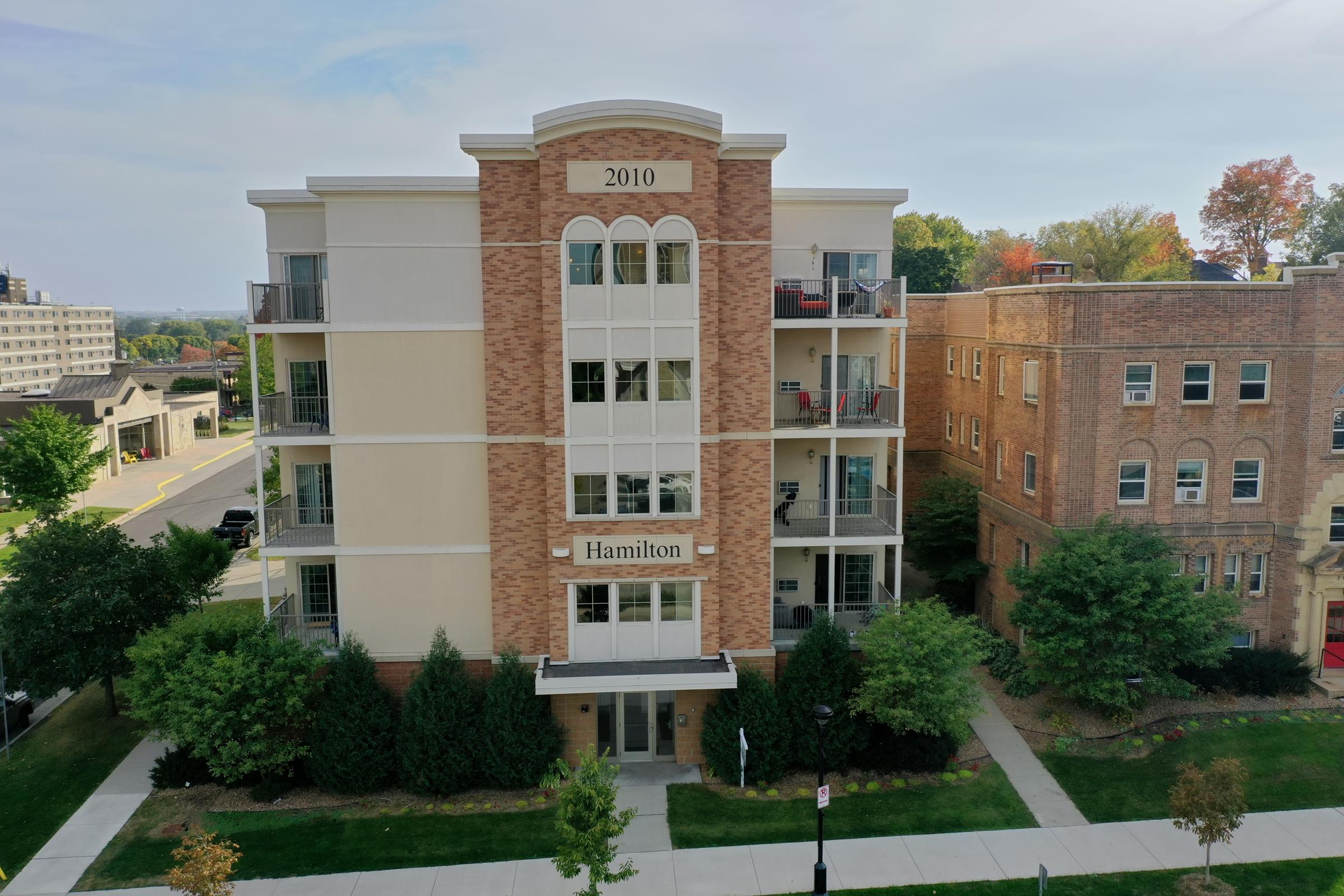 A modern multi-story residential building with the address "2010 Hamilton" prominently displayed. The facade features brick and light-colored panels, with several balconies visible. Surrounding greenery includes trees and shrubs, and the scene is set against a clear sky in an urban environment.
