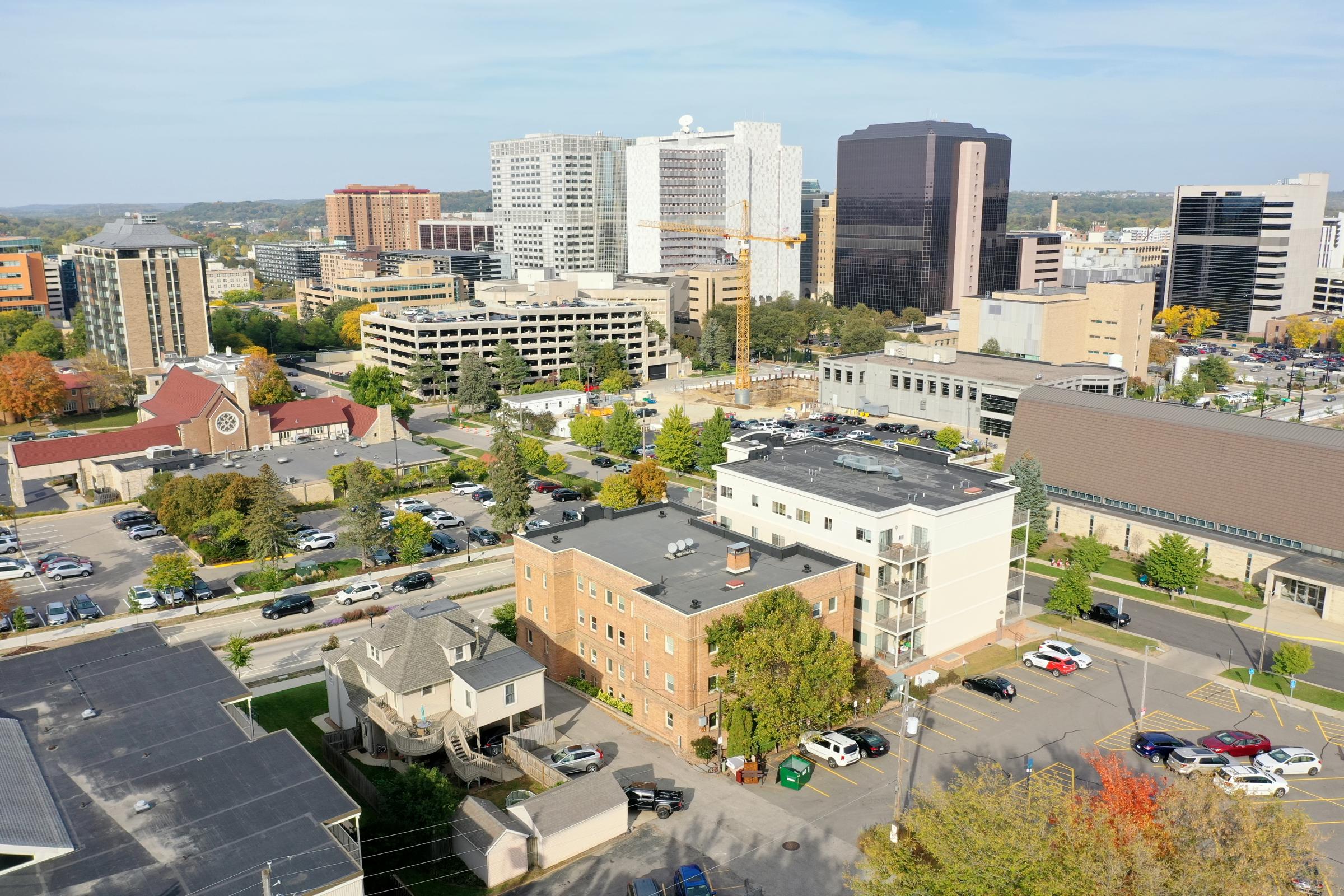 Aerial view of a city skyline featuring a mix of modern and older buildings, parking lots, and trees. Notable construction cranes suggest ongoing development. The foreground shows a brick building alongside a mix of residential and commercial structures, with plenty of parked cars.
