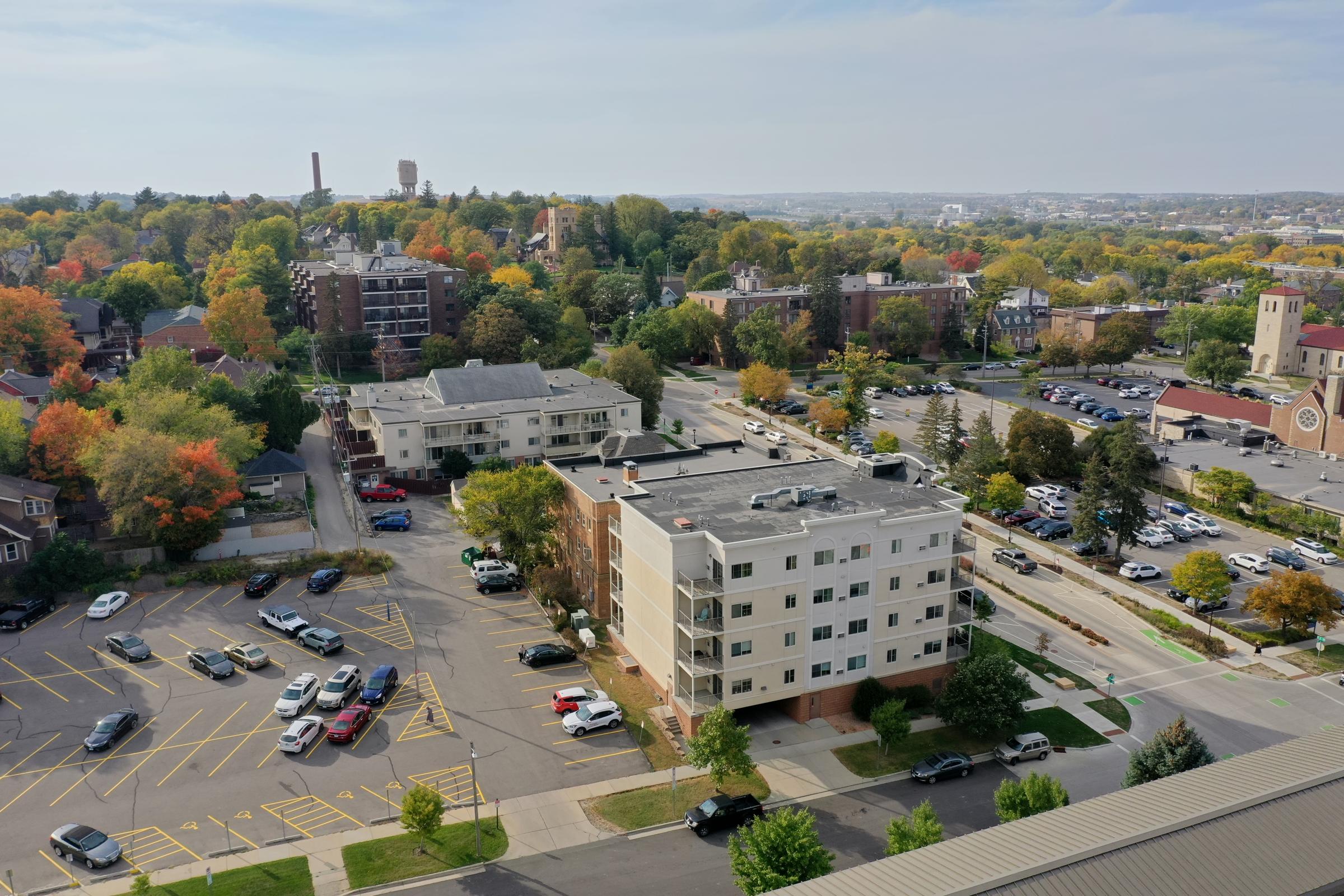 Aerial view of a suburban area featuring a mix of residential buildings and parking lots. Trees with autumn foliage create a colorful landscape. The scene includes a variety of parked cars and distant hills, with a cityscape in the background under a clear sky.