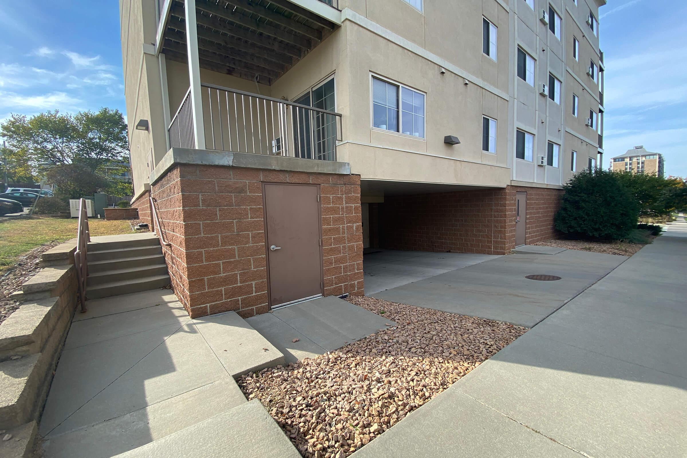 Exterior view of a multi-story building with a covered parking area and stairs leading up to the entrance. The ground features a mix of concrete and gravel, with some greenery in the background. The sky is clear and bright, indicating a sunny day.