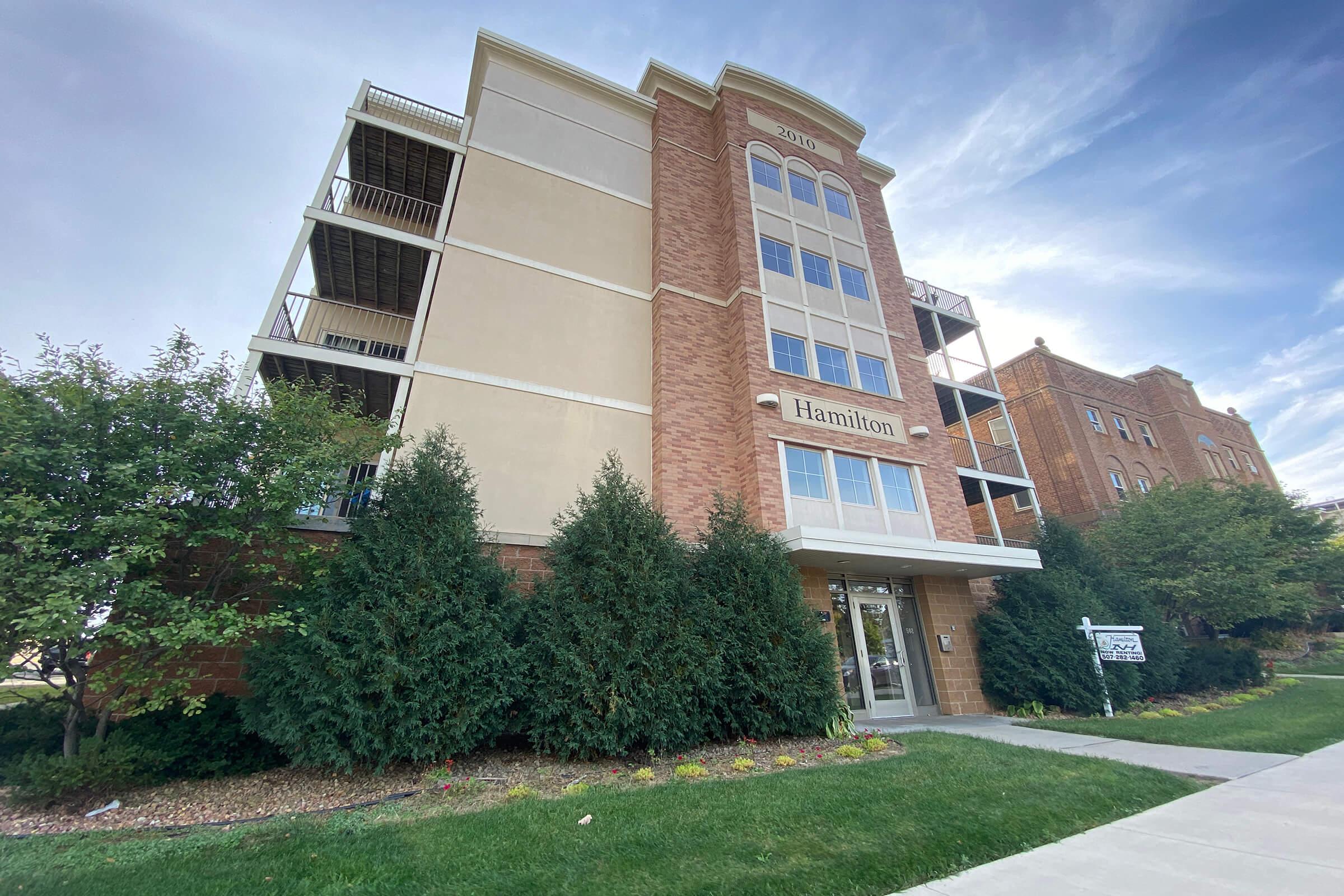 A modern, multi-story building with a brick facade and large windows. The entrance features the name "Hamilton" prominently displayed. Surrounding the building are landscaped bushes and grassy areas, with a walkway leading up to the entrance. The sky is partly cloudy, suggesting a pleasant day.