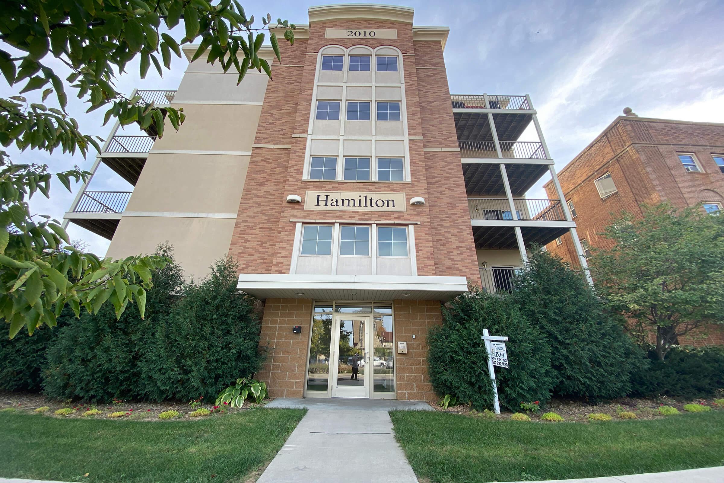 A modern multi-story apartment building with a brick facade. The entrance features a glass door with the name "Hamilton" displayed above. Flanking the entrance are well-maintained shrubs and flowers. The building has balconies and a sign indicating address information.