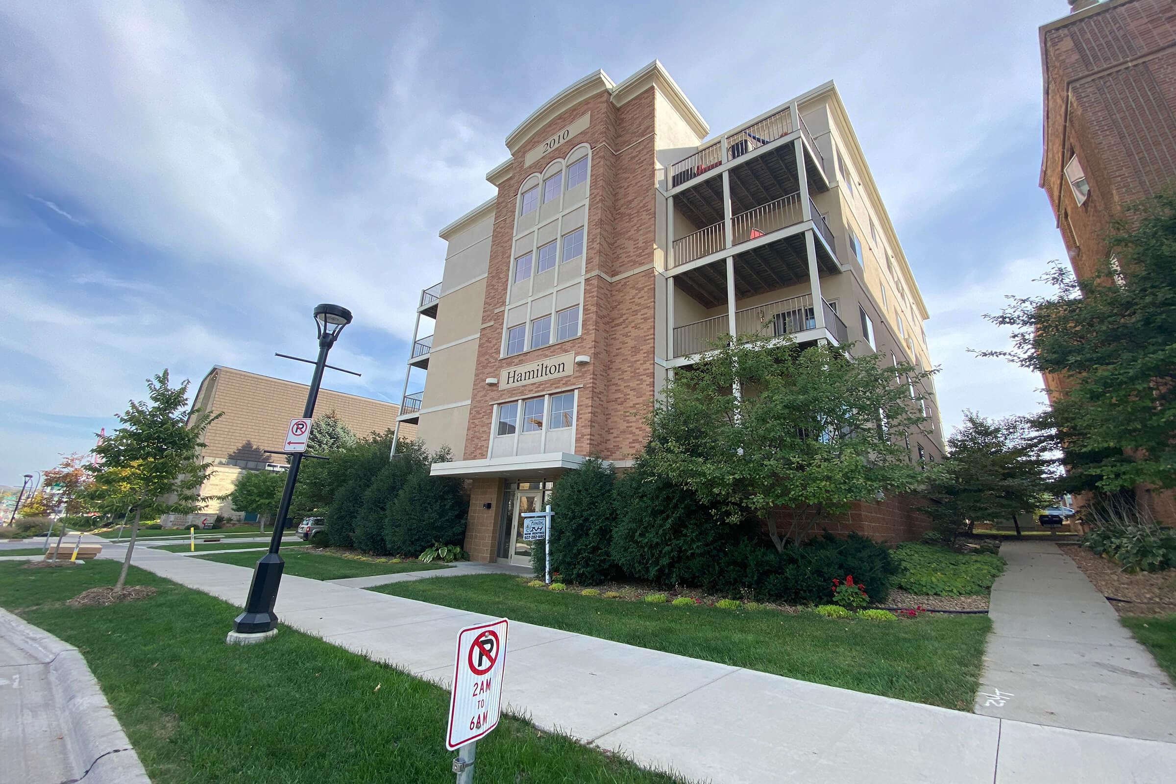 A modern, multi-story residential building with a brick facade and balconies on the upper floors. Lush greenery surrounds the entrance and a street lamp is visible in the foreground, along with a no parking sign. The sky is partly cloudy, indicating clear weather.