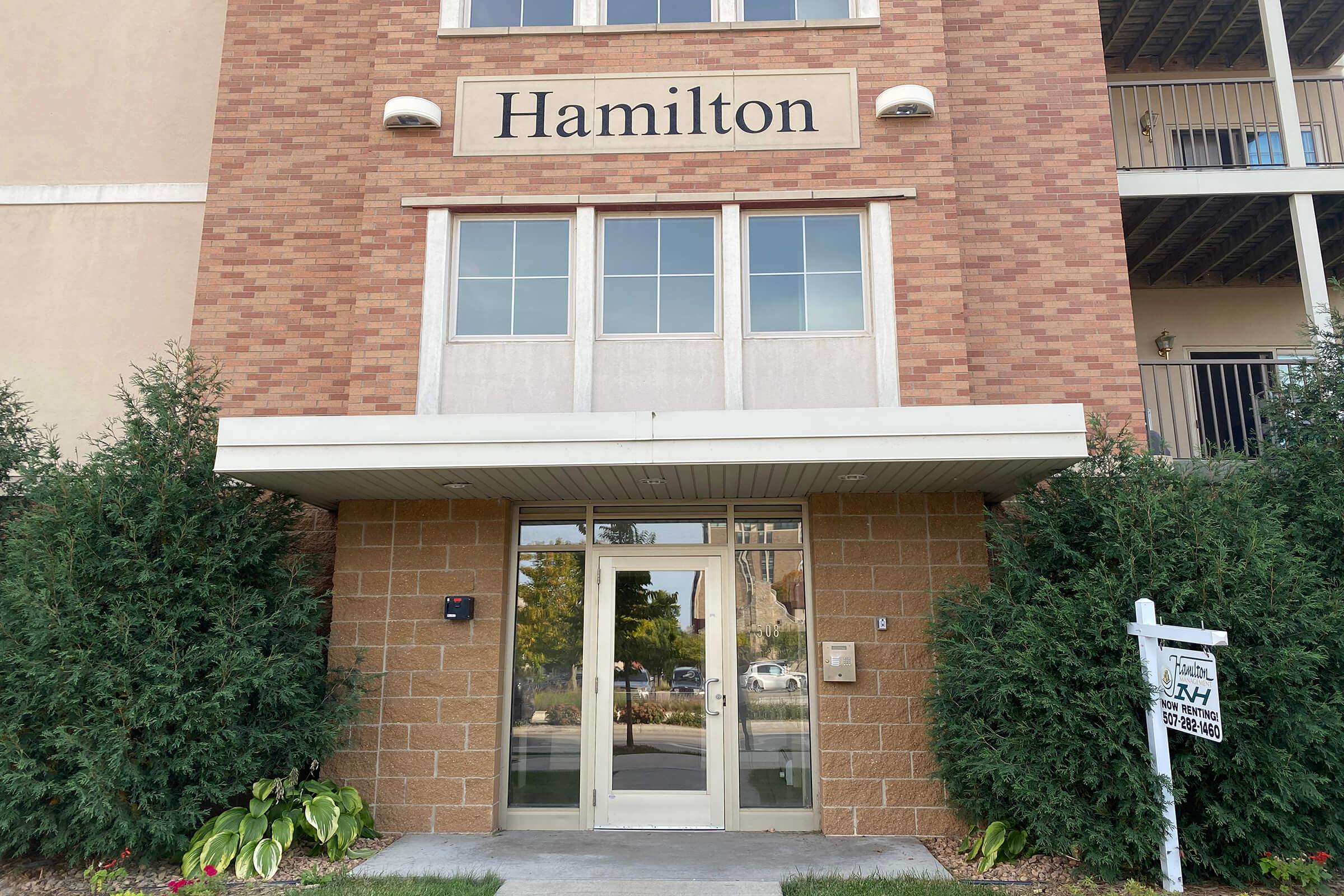Entrance of a building with the name "Hamilton" displayed prominently above the door. The facade features brick and cream-colored siding, with two windows on the upper level. Flanking the entrance are green shrubs and a for-sale sign nearby.