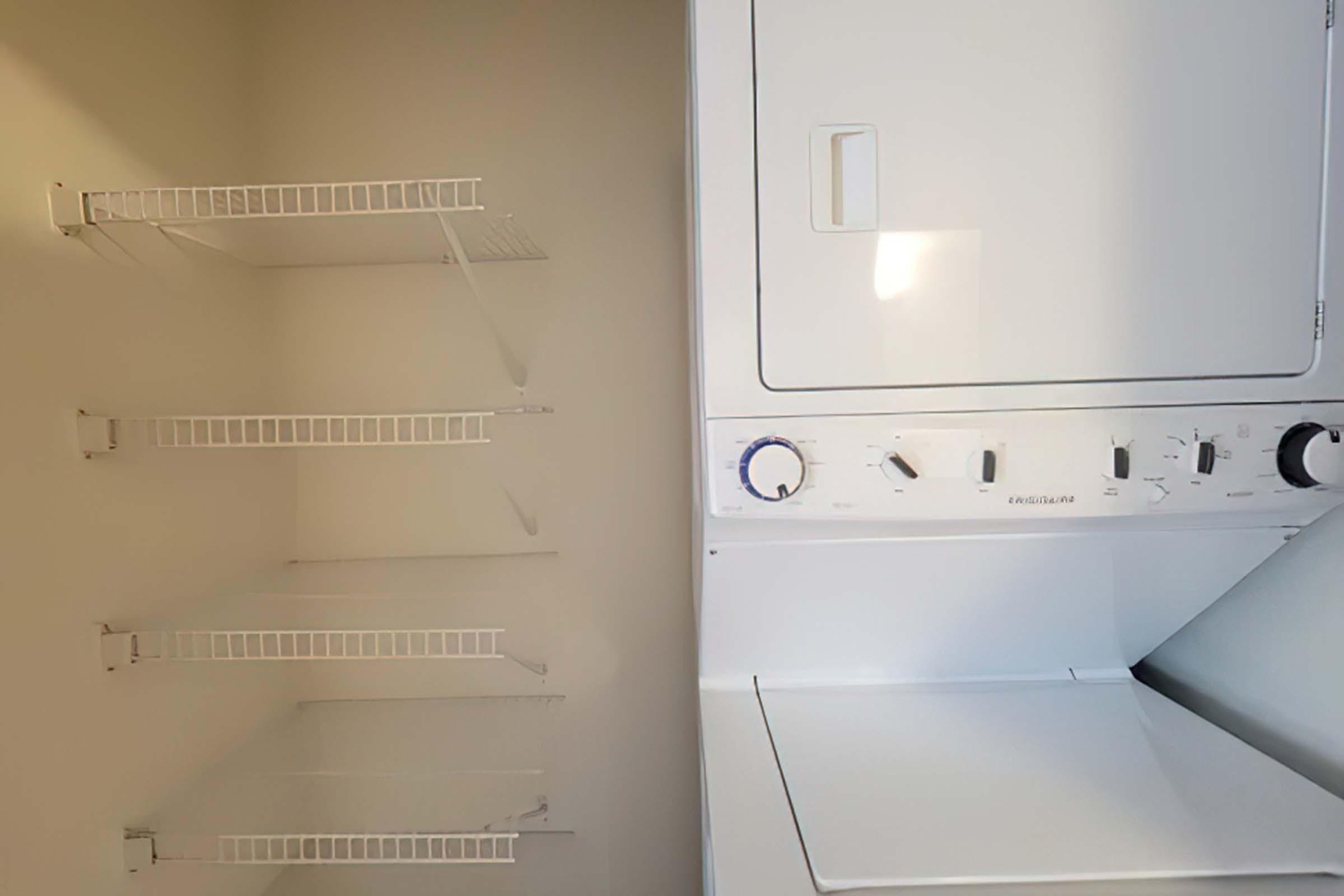 A laundry area featuring a stacked washer and dryer on the right side, with three empty wire shelves on the left side. The walls are a neutral color, and there is natural light illuminating the space.