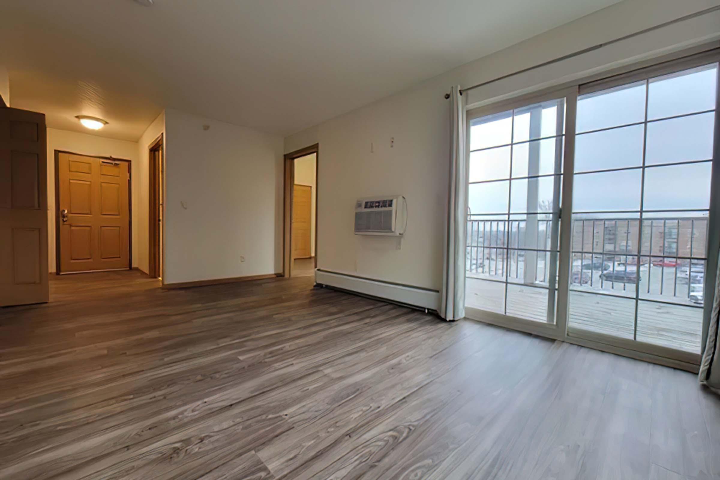A spacious, empty room with wooden flooring, featuring a large window with a sliding door that leads to a balcony. The walls are painted light, and there's an air conditioning unit on one wall. In the background, a doorway leads to another room and a main entrance door is visible.
