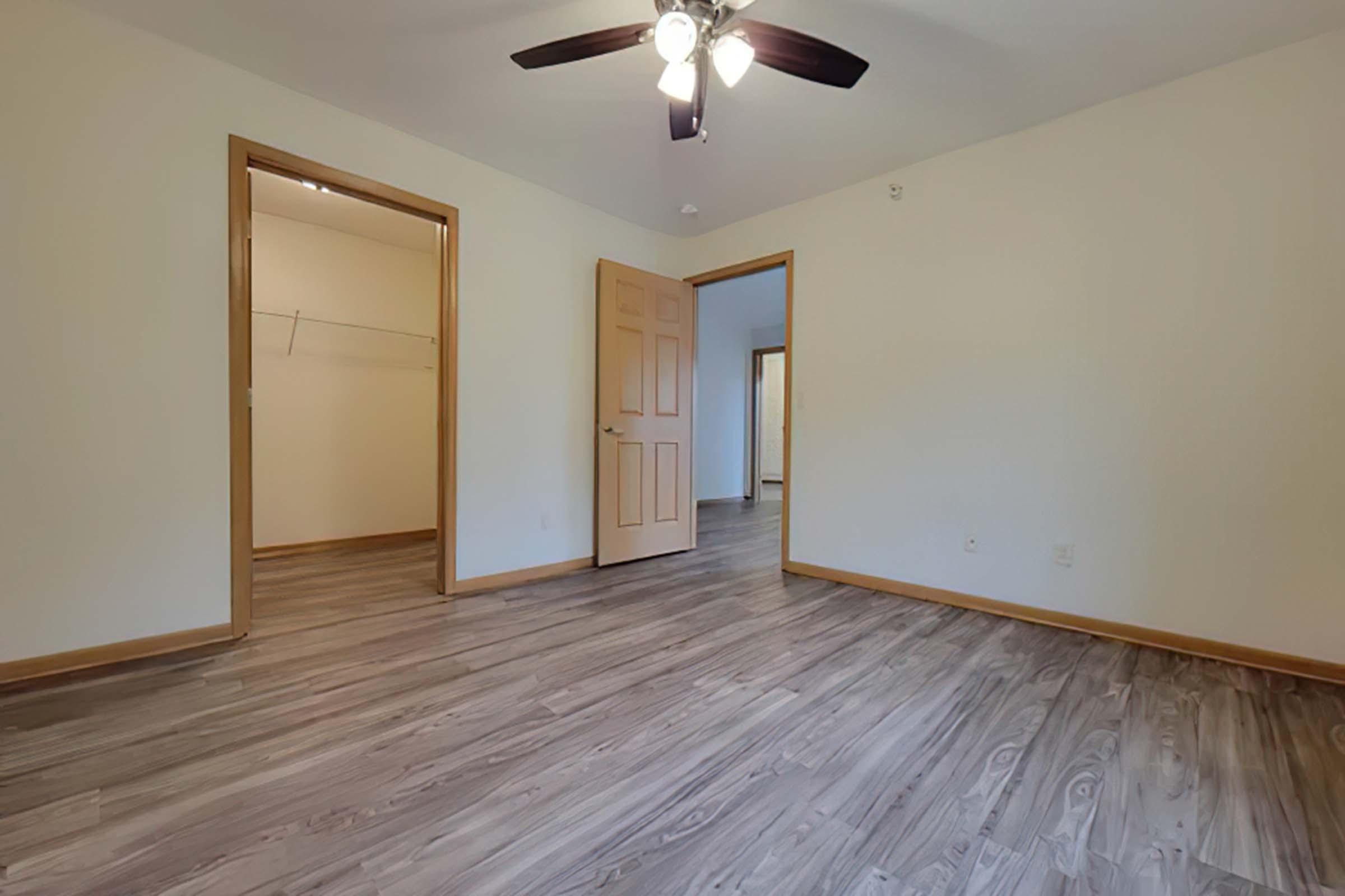 A spacious, empty room featuring light-colored walls and a ceiling fan. Two doorways lead to other areas, and there's a closet visible on the left. The flooring is wood-like and stretches throughout the room, creating a clean and modern appearance.