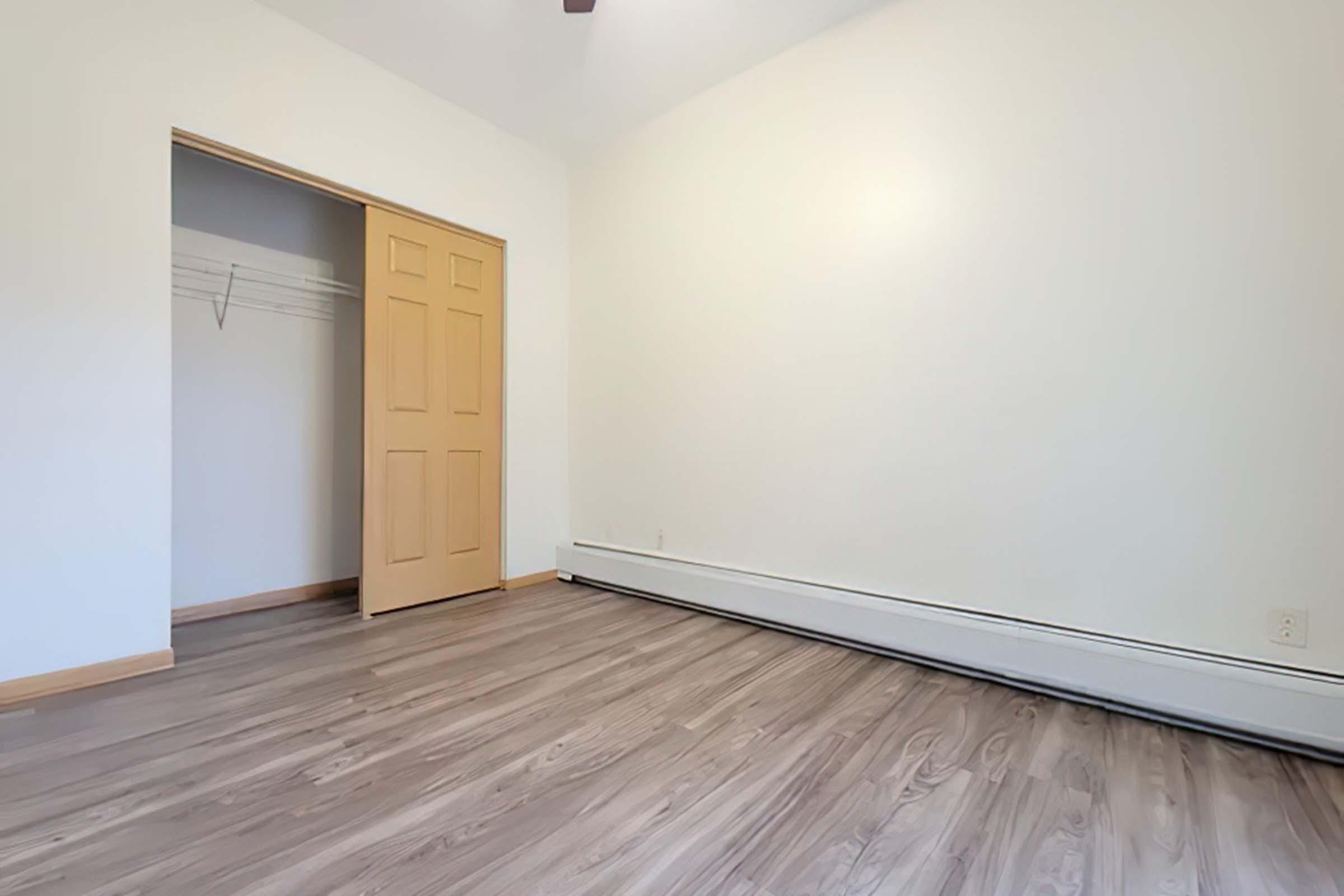 A spacious, empty room with wooden flooring and light-colored walls. There is a closet with double doors on the left side, and the room has a ceiling fan. The overall ambiance is bright and minimalistic, suggesting potential for various uses or decoration.
