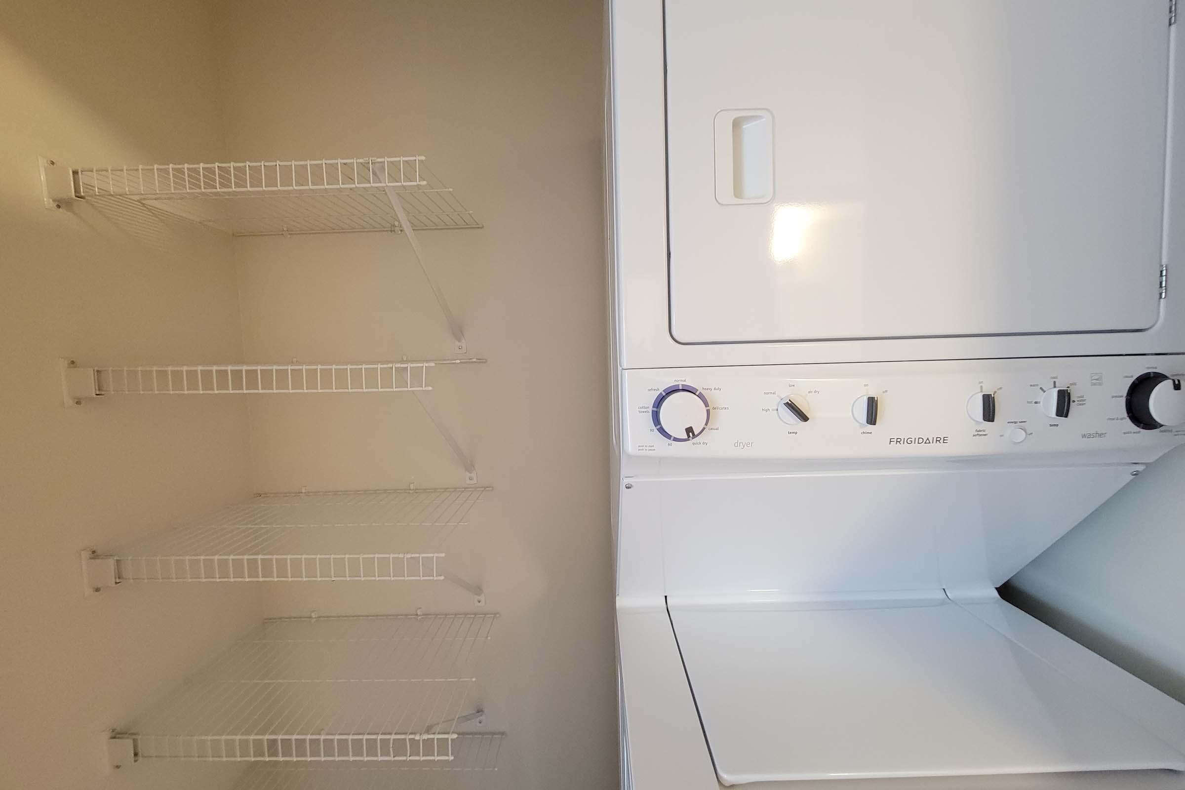 A laundry area featuring a white washer and dryer unit, with a control panel on the front. Adjacent to the appliances are three empty wire shelves mounted on the wall, providing storage space for laundry supplies. The walls are painted a neutral color.