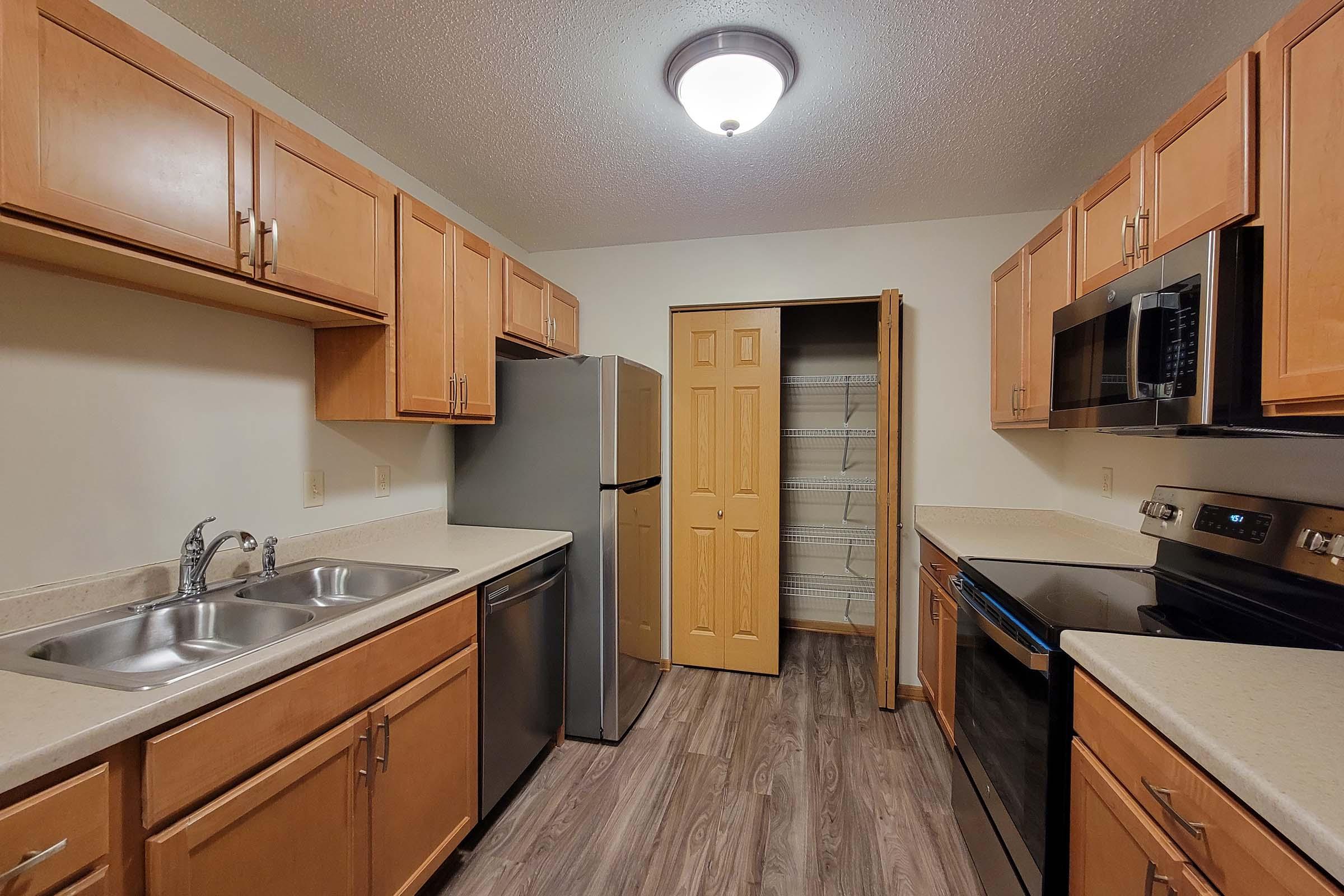 Modern kitchen featuring oak cabinets, stainless steel appliances including a refrigerator and oven, a double sink, and a countertop. The kitchen has laminate flooring and a pantry with bi-fold doors. A ceiling light fixture illuminates the space.
