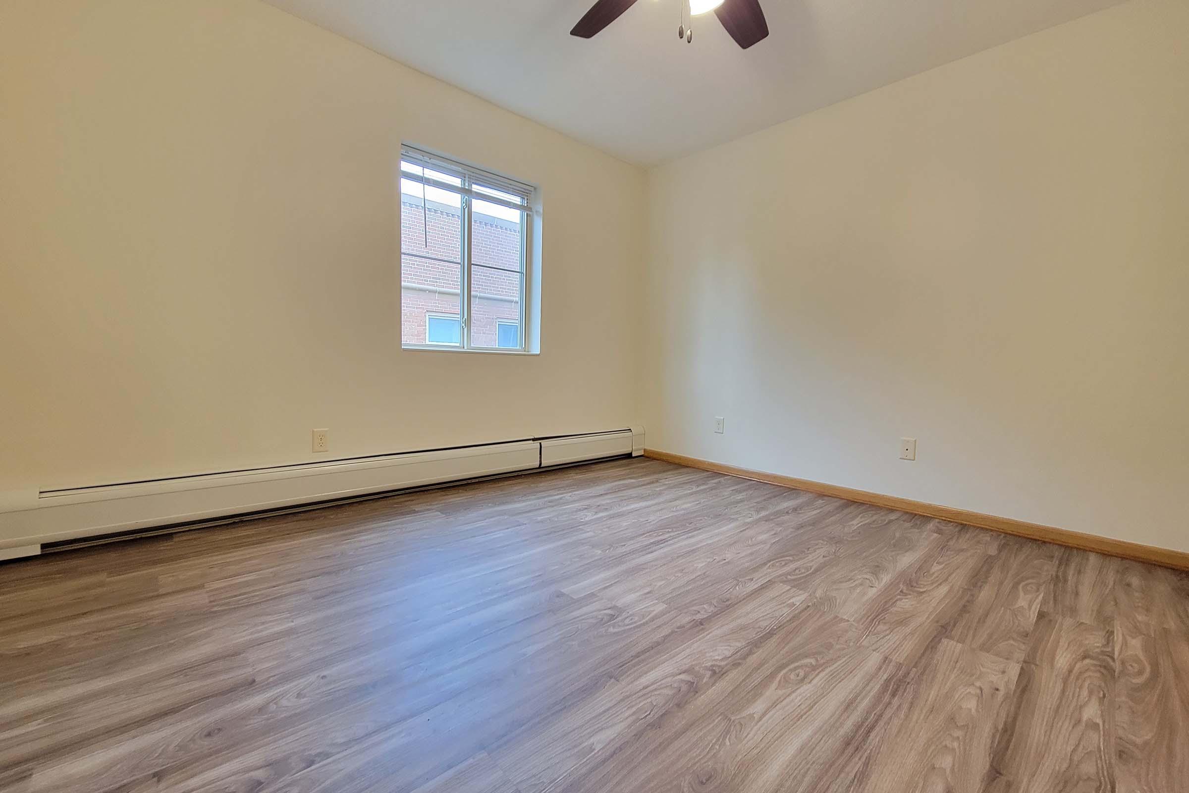 Empty room with light-colored walls and a wooden floor. A ceiling fan is visible, and there are two small windows on one wall providing natural light. The overall atmosphere is bright and spacious, suitable for various uses.