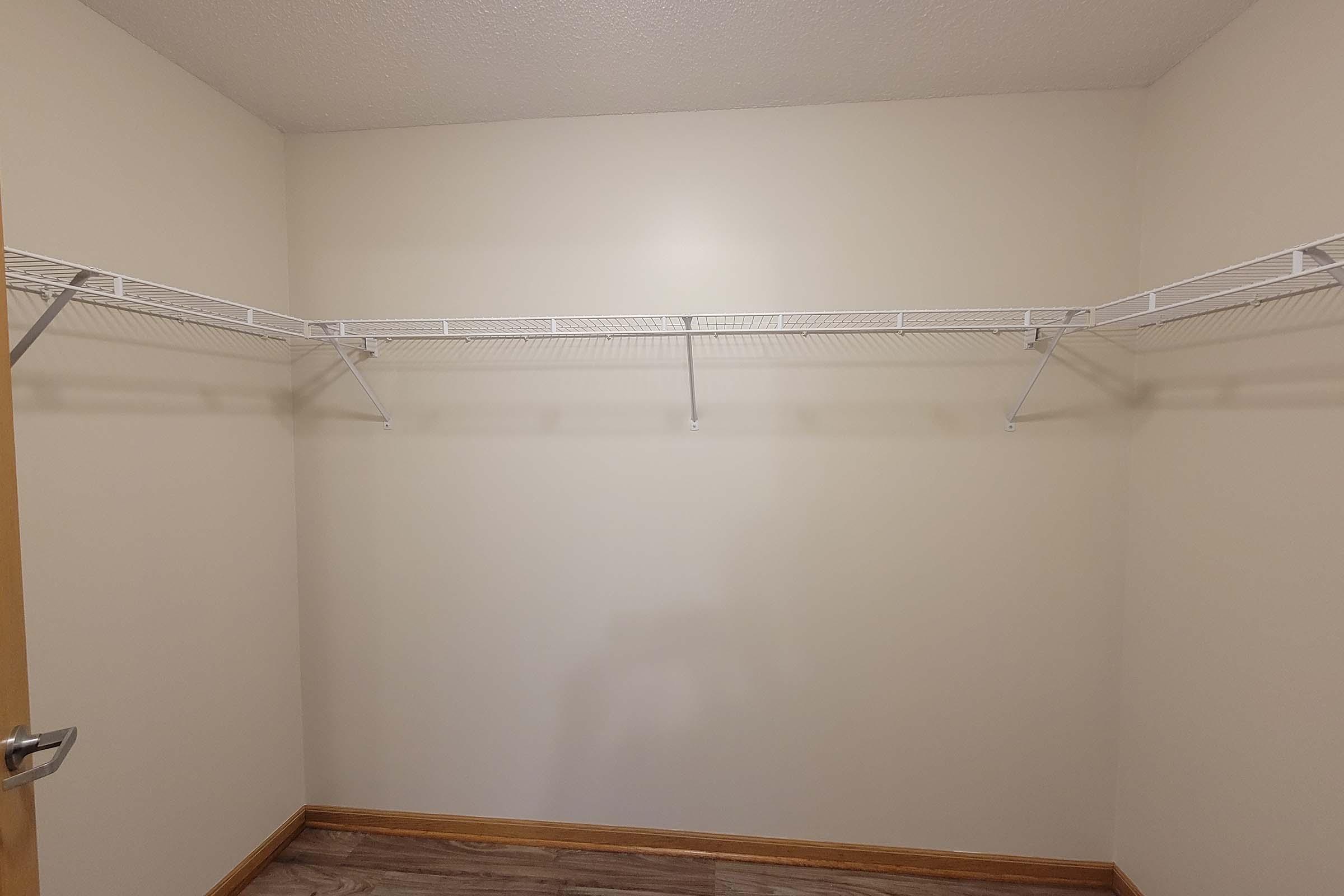 Empty closet with light-colored walls and a wooden floor. There are three wire shelves mounted on the wall, with space beneath them. A door handle is visible on the left side. The overall space appears bright and minimalistic.