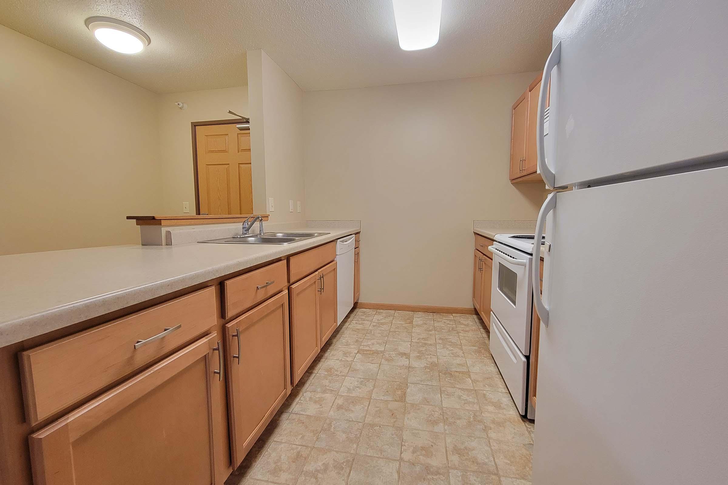 A view of a kitchen featuring light wood cabinets, a white refrigerator, an oven, and a sink. The room is bright with neutral-colored walls and a tiled floor. There is a doorway leading to another room, and the overall space appears clean and functional.