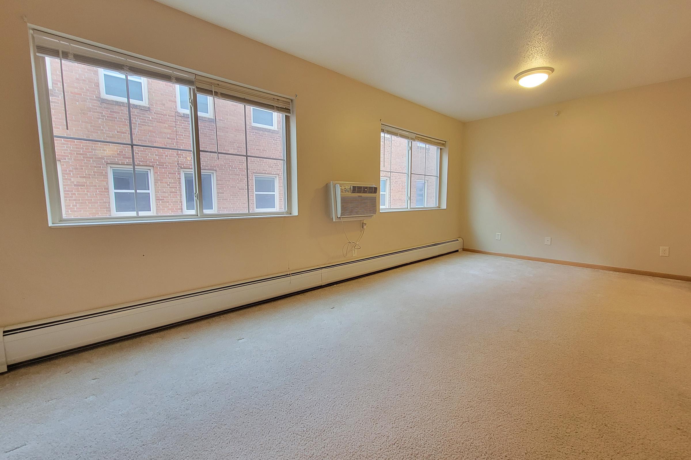 A spacious, empty room with light beige walls and carpet. Two large windows allow natural light, revealing a view of a brick wall outside. An air conditioning unit is mounted on the wall, providing comfort. The overall atmosphere is bright and airy, with minimal furnishings.