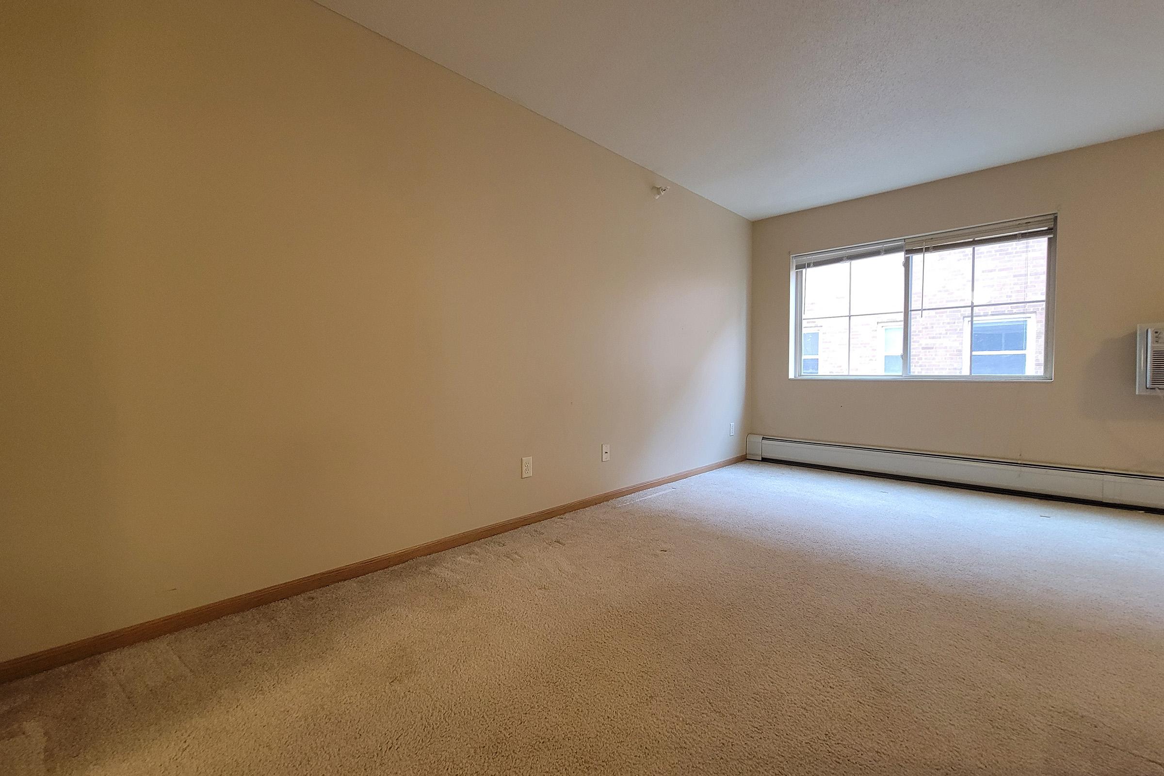 Empty room with beige walls and light beige carpet. There is a window on the far wall with a view of the outside. An air conditioning unit is visible under the window. The space is well-lit with natural light, creating a bright and open ambiance.