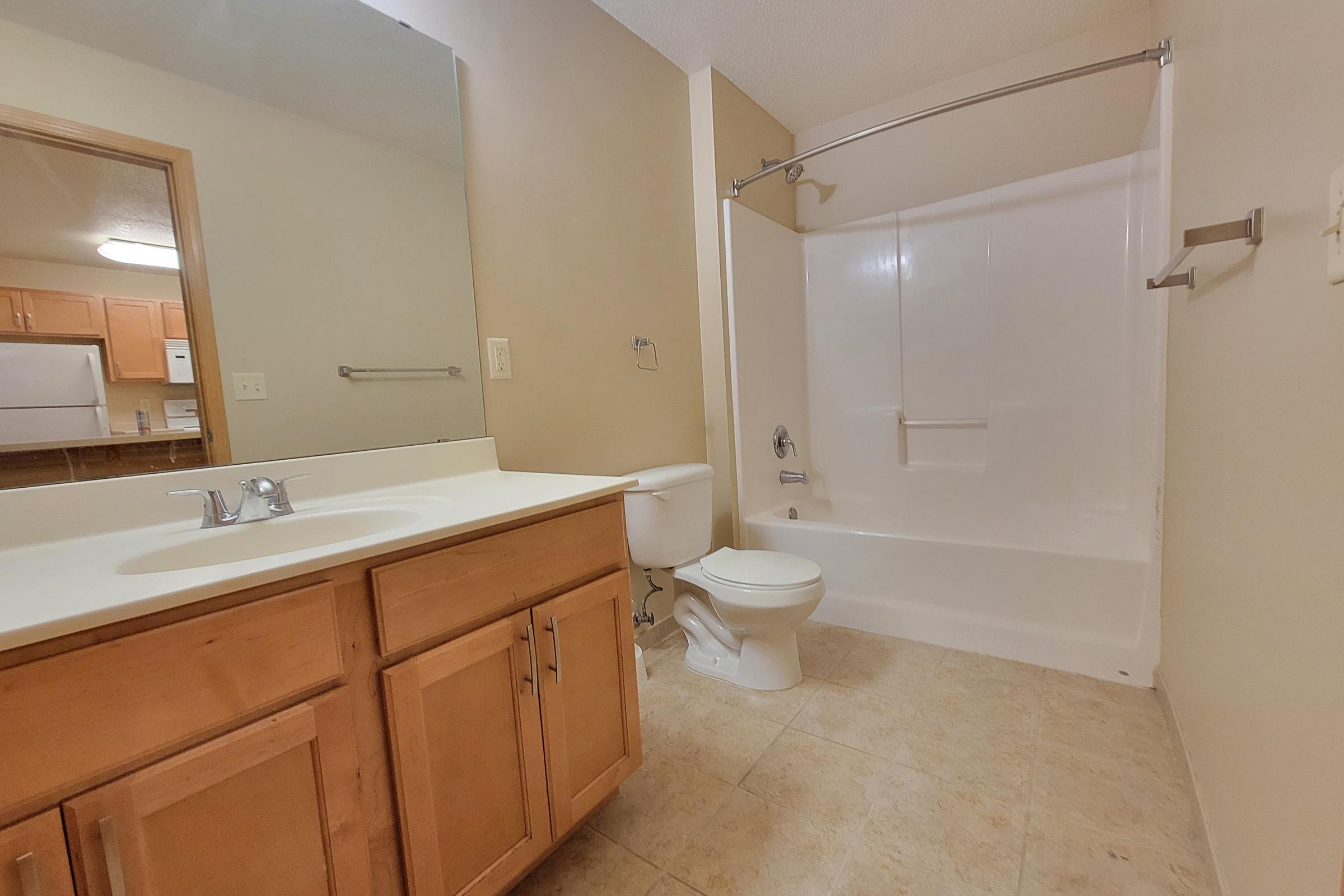A clean, well-lit bathroom featuring a wooden vanity with a sink, a toilet, and a tub-shower combination. The walls are painted in a neutral color, and the floor is tiled. In the background, there is a glimpse of a kitchen area.