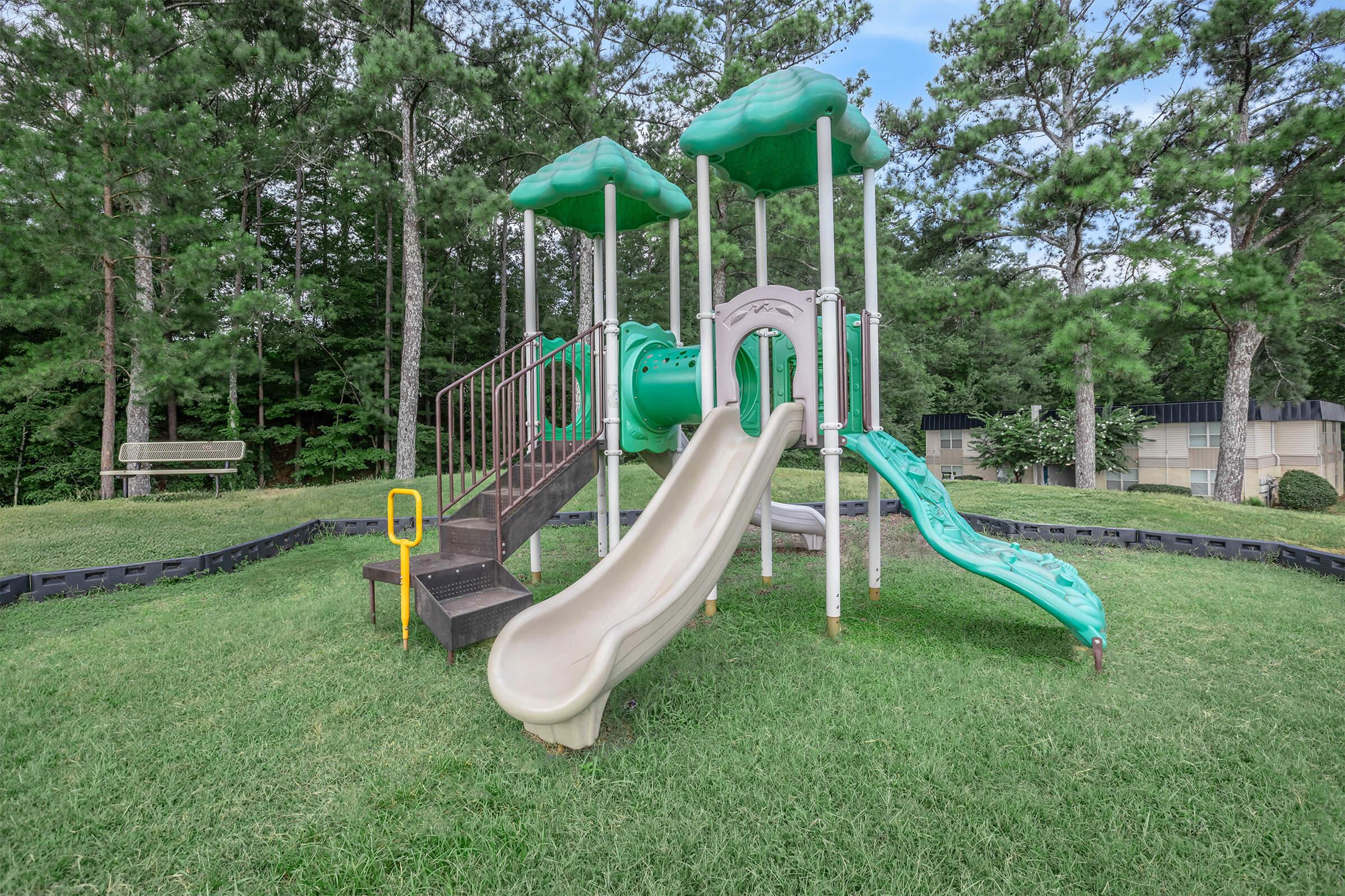Playground equipment featuring a green slide structure with two climbing sections and a tunnel, surrounded by grassy areas and trees in the background. The area appears spacious and well-maintained, ideal for children to play and explore.